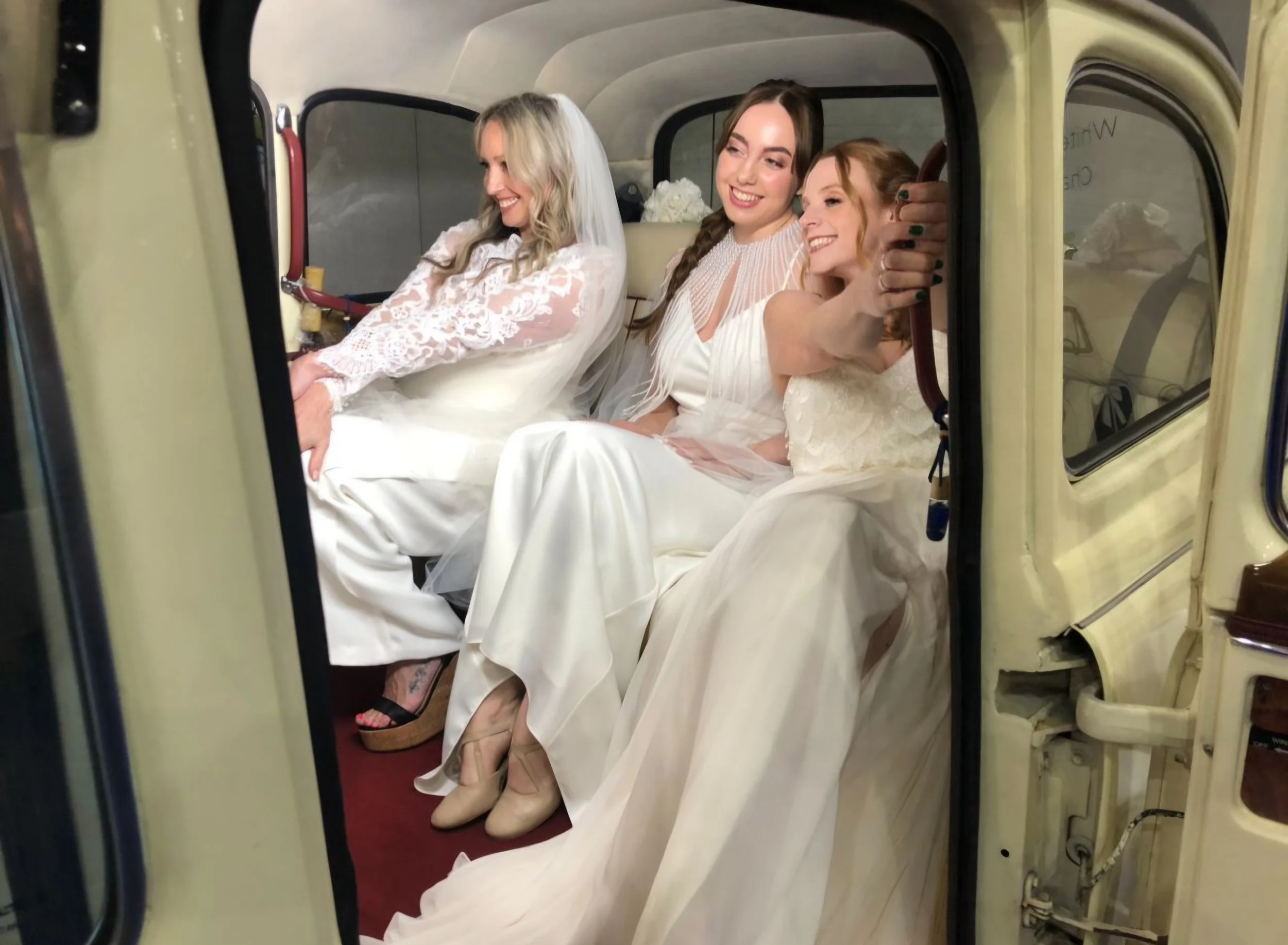 Three women in white wedding dresses sitting inside a vintage vehicle, smiling, with one woman showing her toenails and holding hands together.