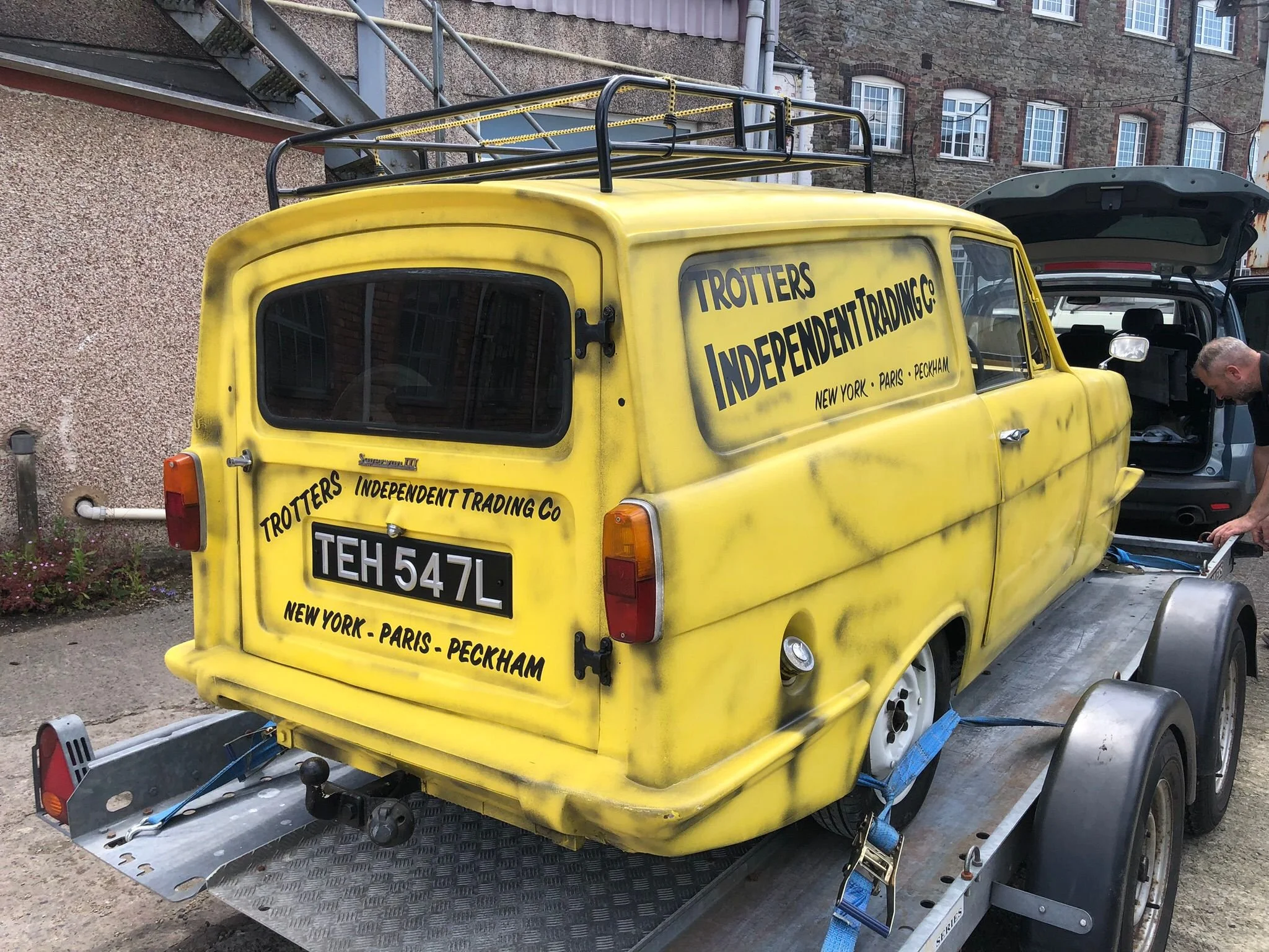 A yellow utility vehicle with signage for Trotters Independent Trading Co, with locations in New York, Paris, Peckham. The vehicle is on a trailer or flatbed, with a man working beside it, possibly repairing or inspecting. The background has a brick building with multiple windows.