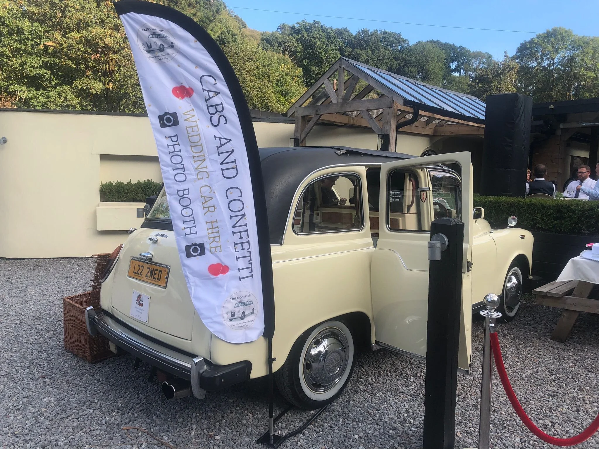 A vintage cream-colored car parked outdoors with a banner in front of it that reads 'Cotswold Car Hire' and features images of classic cars. The car is situated on gravel, with a building and a group of people sitting and talking in the background.