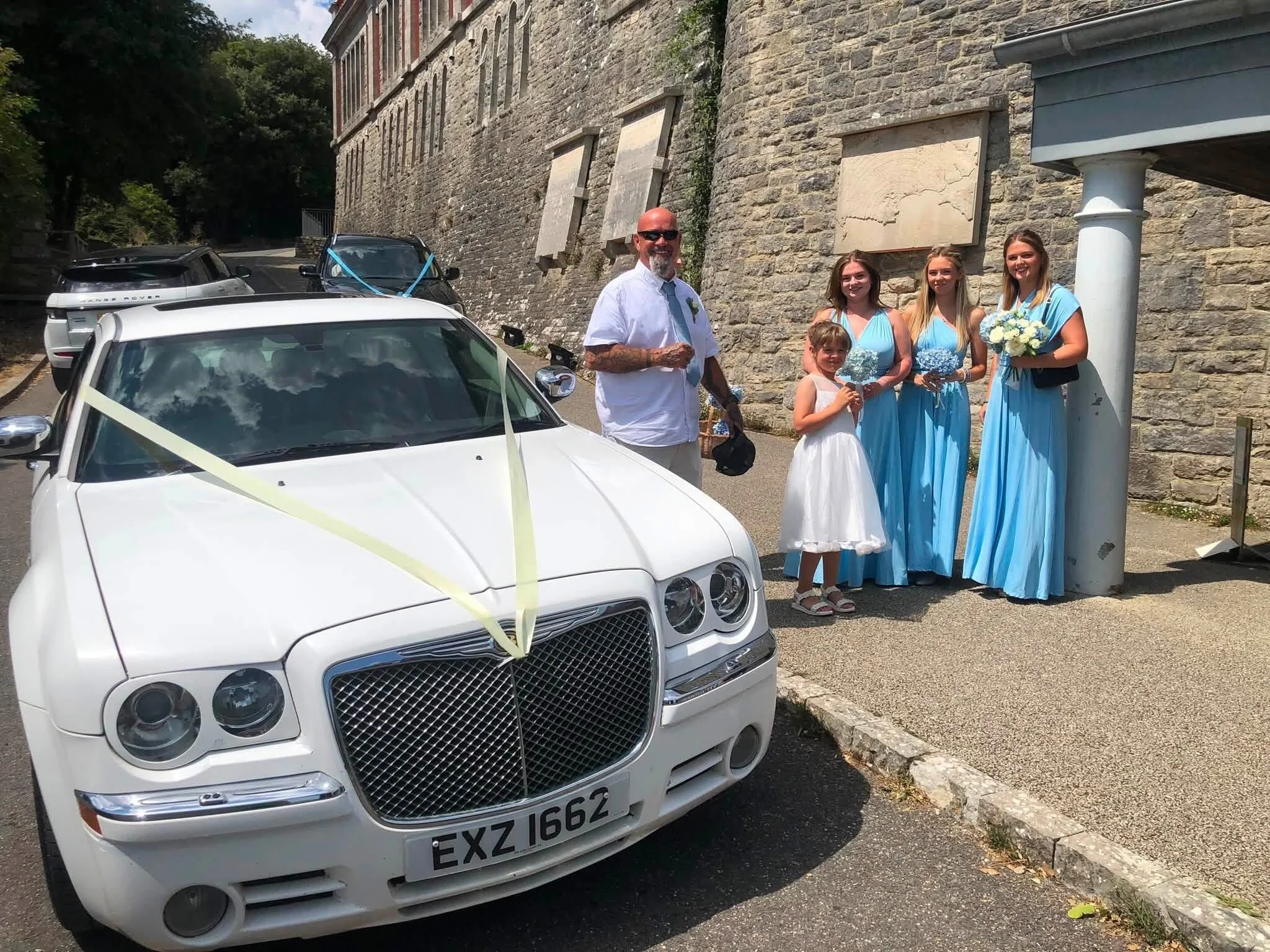 Wedding scene with a group of four women in blue dresses, a girl in white holding flowers, and a man with glasses, white shirt, and tie standing near a decorated white car with a yellow ribbon.