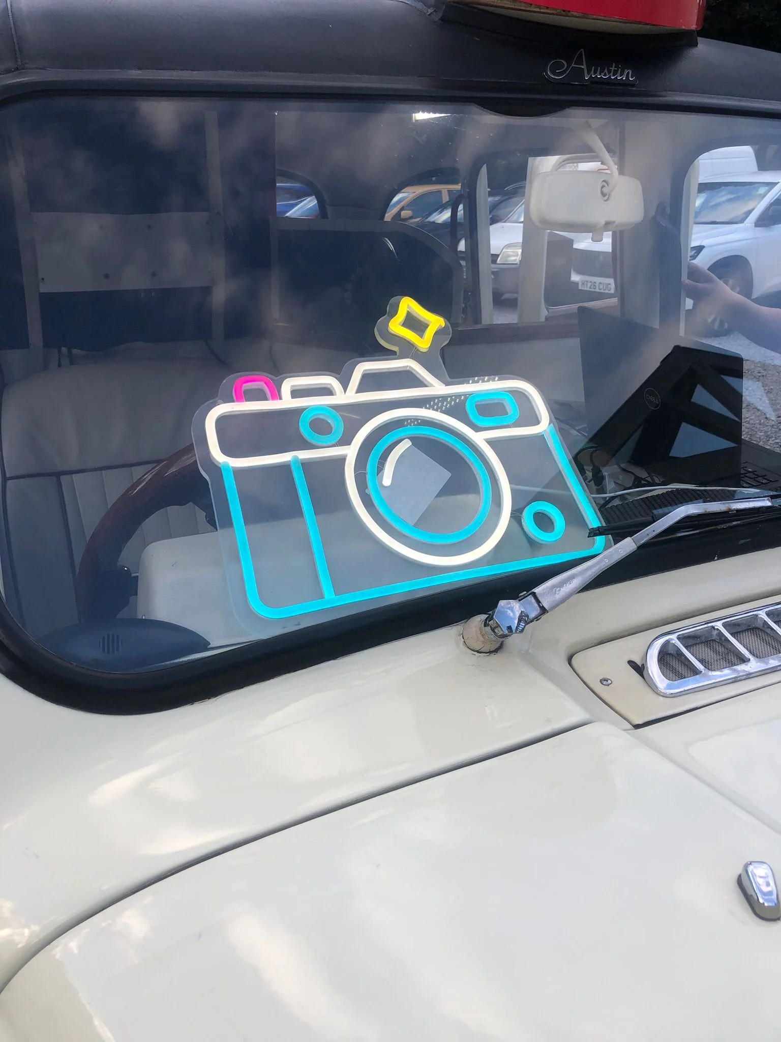 Reflective surface on a car windshield showing a colorful neon sign of a camera with pink, blue, and yellow elements, and some parking lot and cars in the background.