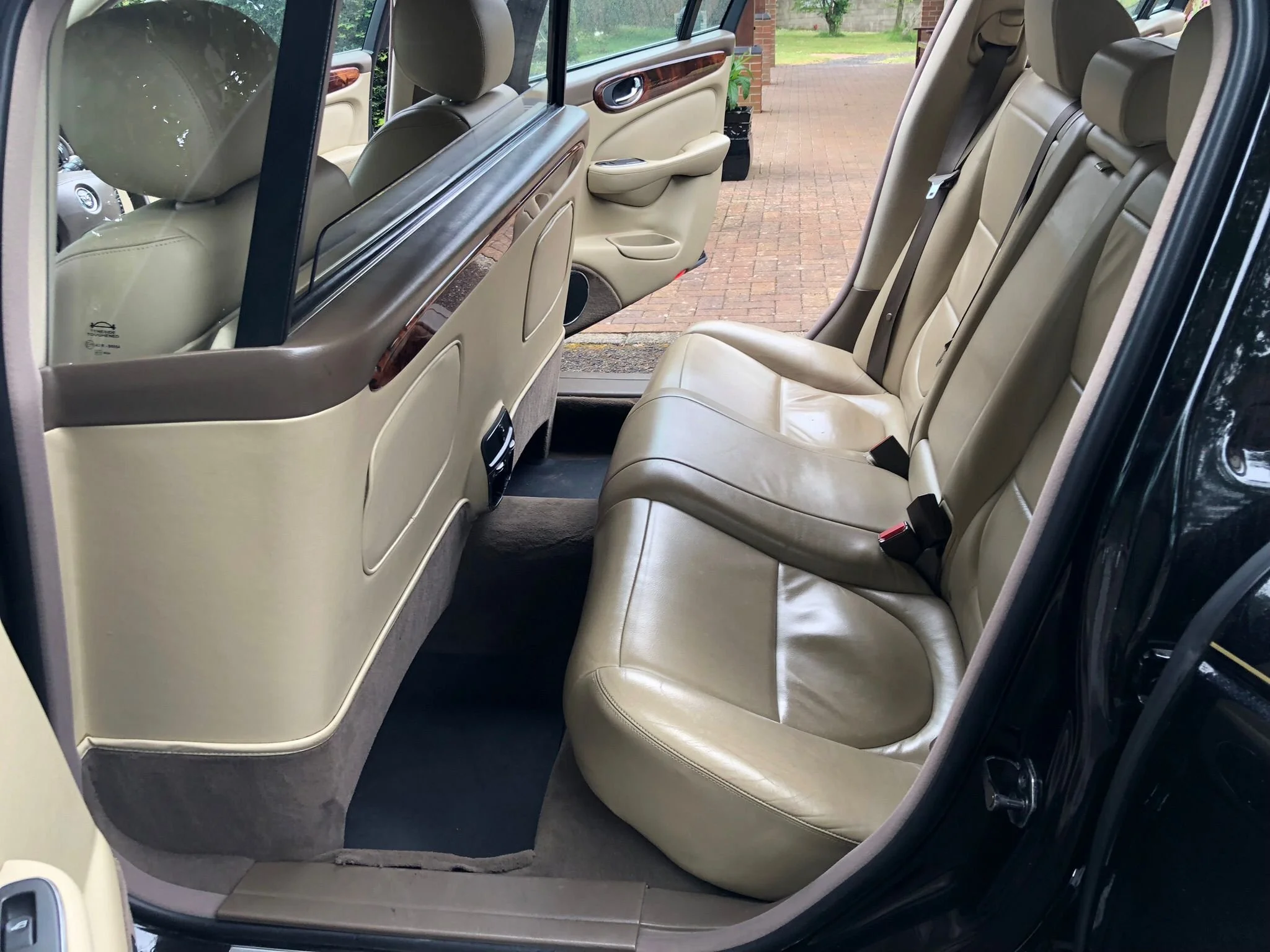 The image shows the interior of a car focusing on the backseat and the rear side door, with beige leather seats and wood trim details on the door panel. The car is parked outside on a brick driveway with greenery visible through the window.