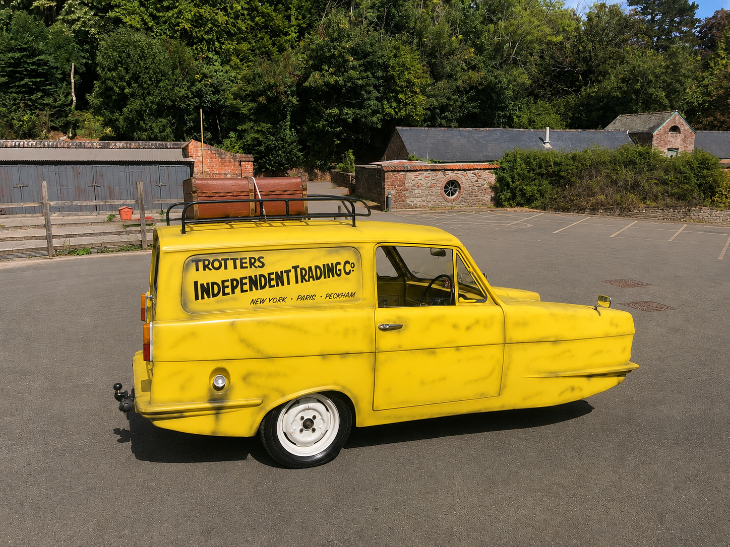 A small yellow vehicle with the words 'Trotters Independent Trading Co.' and locations 'New York, Paris, Peckham' on the side, parked in an empty lot with trees and brick buildings in the background.