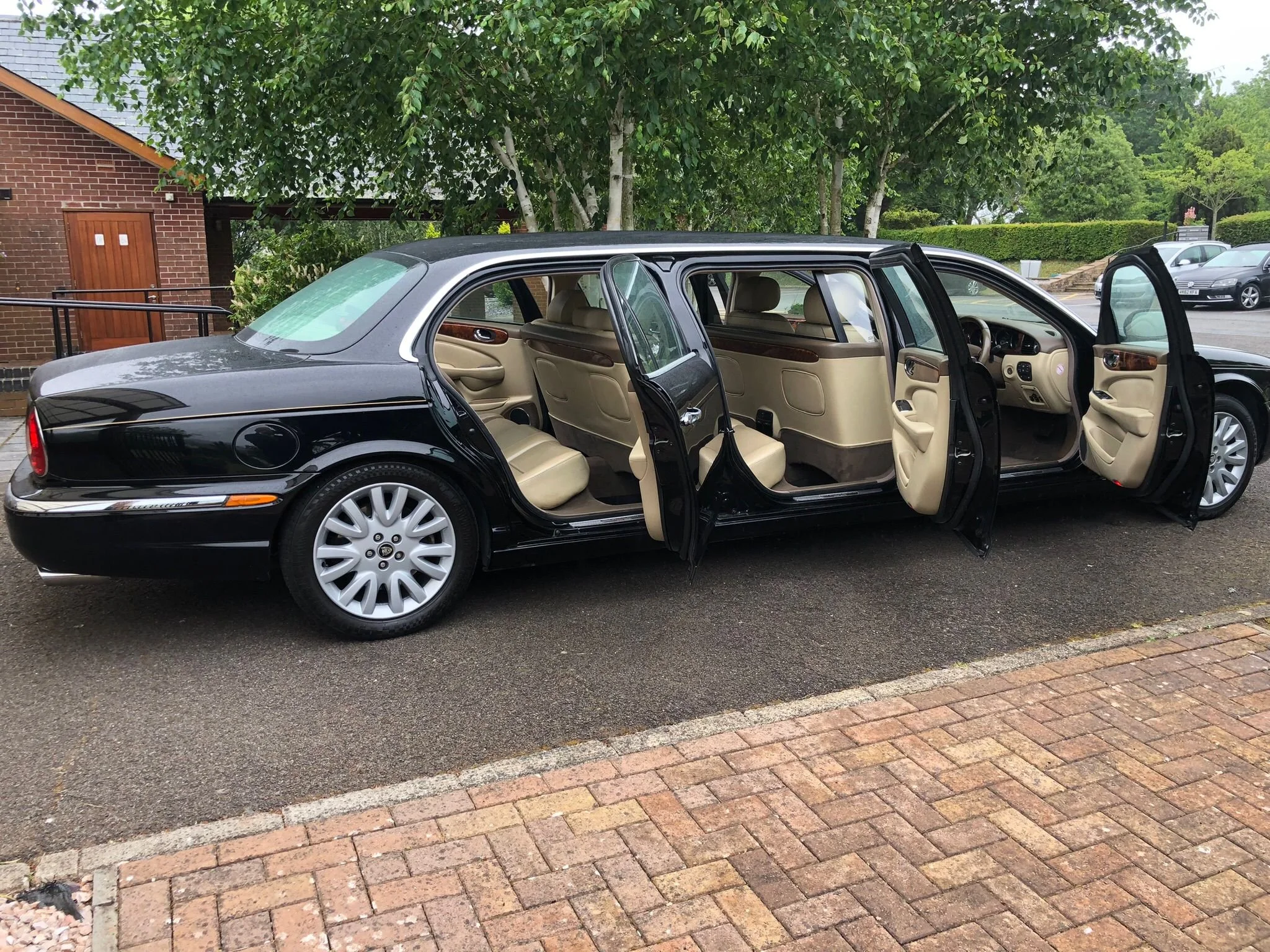A black luxury car with beige interior doors and seats is parked on the street with the rear and front doors open. Green trees and other parked cars are visible in the background.