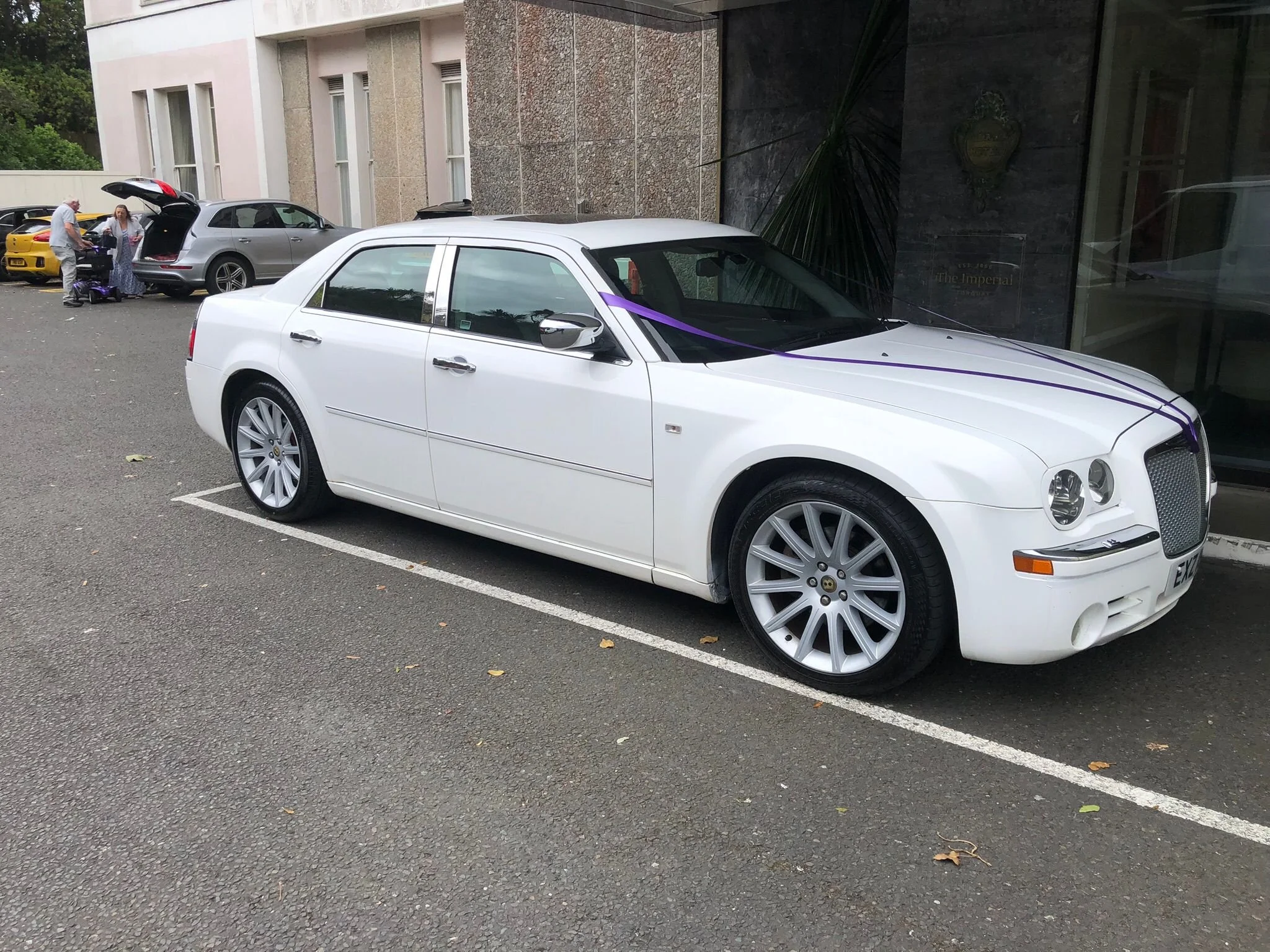 A white Bentley parked in a parking lot in front of a concrete building. The car has purple ribbons on it, indicating it may be part of a celebration or special event.