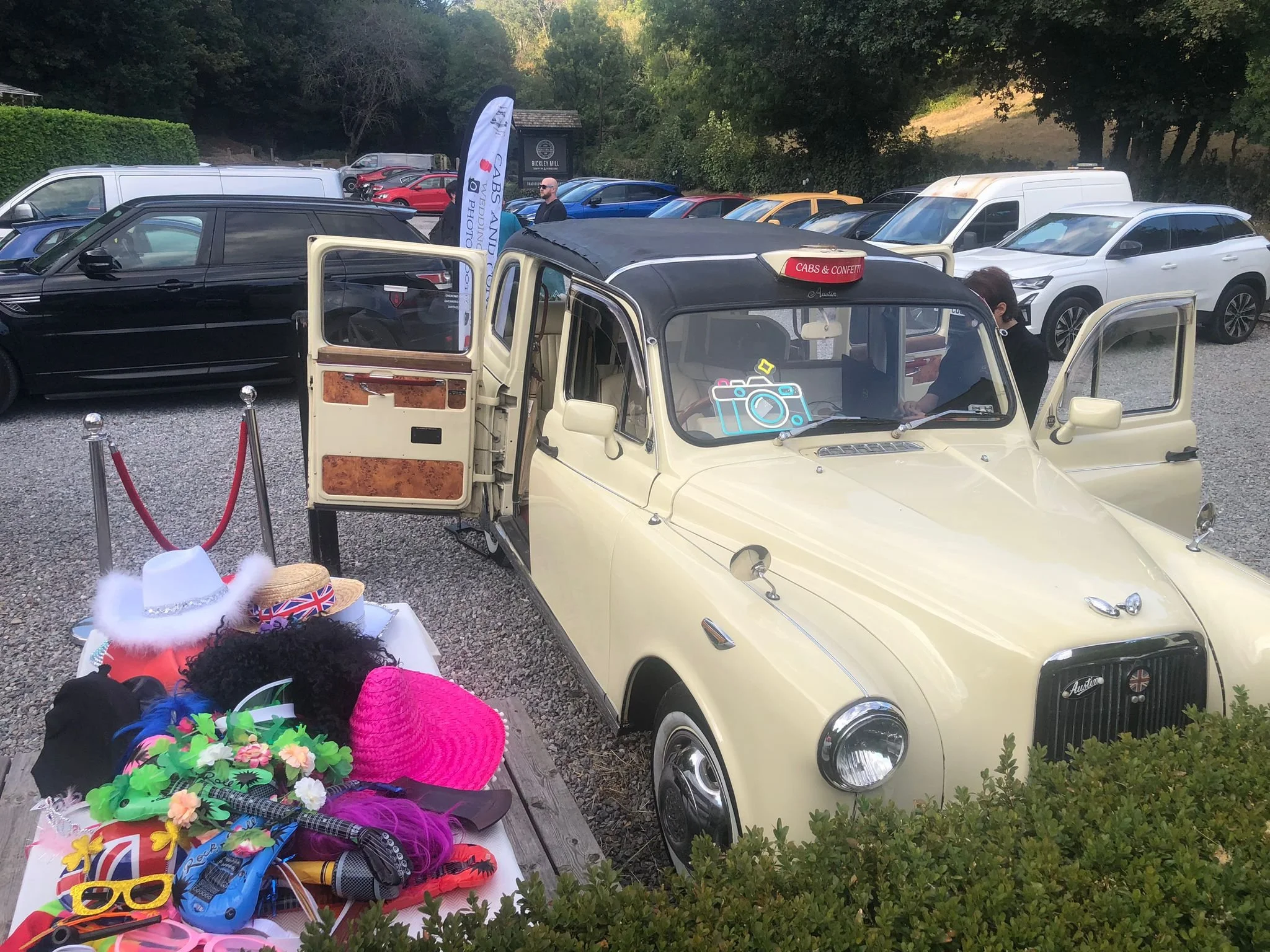 Vintage cream-colored Mini Cooper with open doors parked outdoors at a car gathering. In the background, there are several modern cars, people, and a sign for Buckley Mill. In the foreground, there's a table with colorful hats, headbands, and accessories.