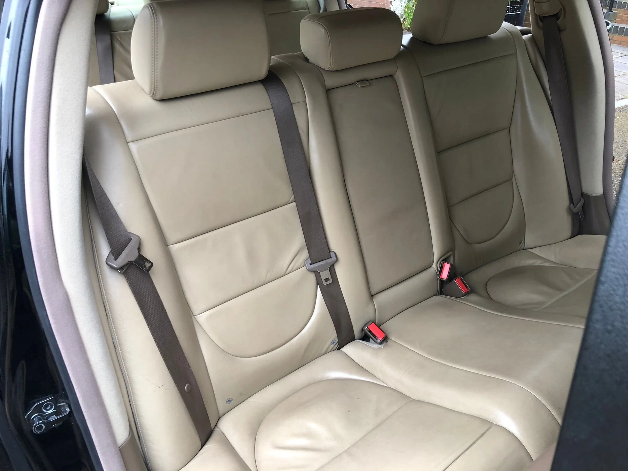 Rear interior of a beige leather car backseat with three headrests and seat belts.