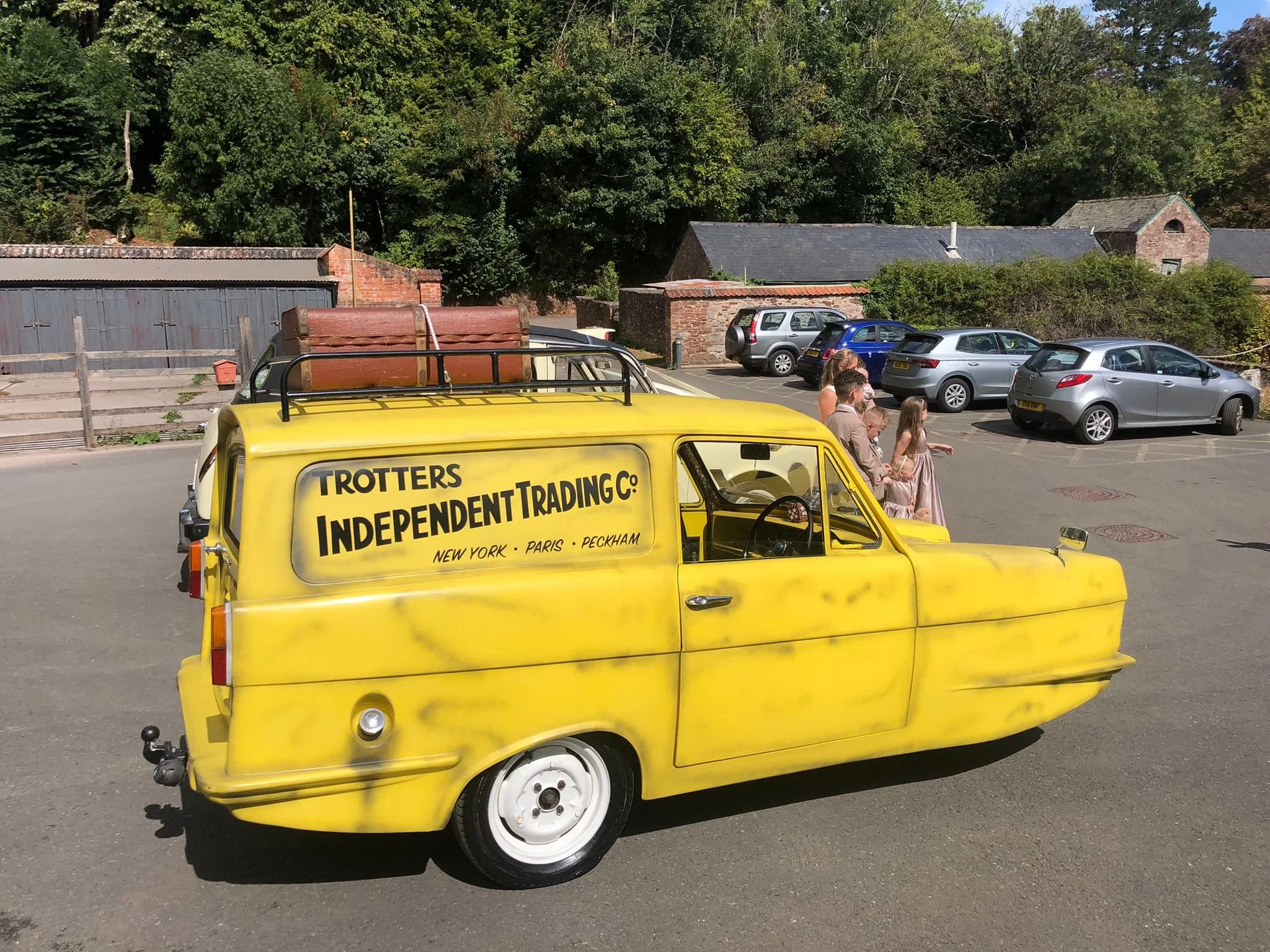 A yellow tuk-tuk vehicle with advertising for independent trading company, parked in a lot with five cars, three women, and group of children, in front of a backdrop of green trees and old brick buildings.