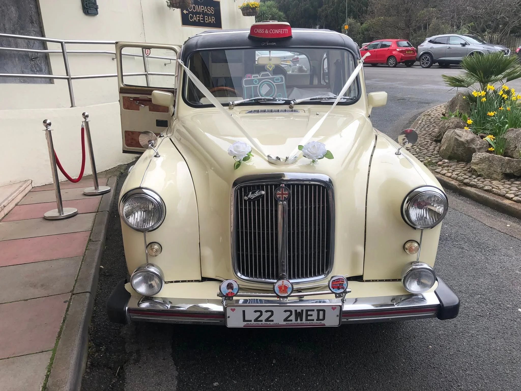 Vintage cream-colored taxi with a red sign on top that says "Cabs & Confetti". The front of the taxi has decorative white ribbons and potted white roses, indicating it may be used for a wedding or special occasion. The license plate reads "L22 2WED" and there are various small decorative items on the front bumper.