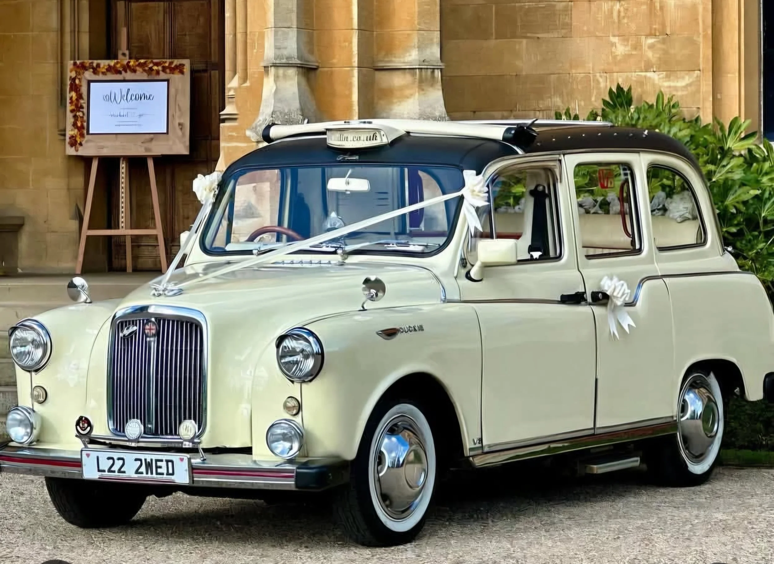 A vintage cream-colored car decorated with wedding ribbons and bows, parked in front of a stone building with a welcome sign.