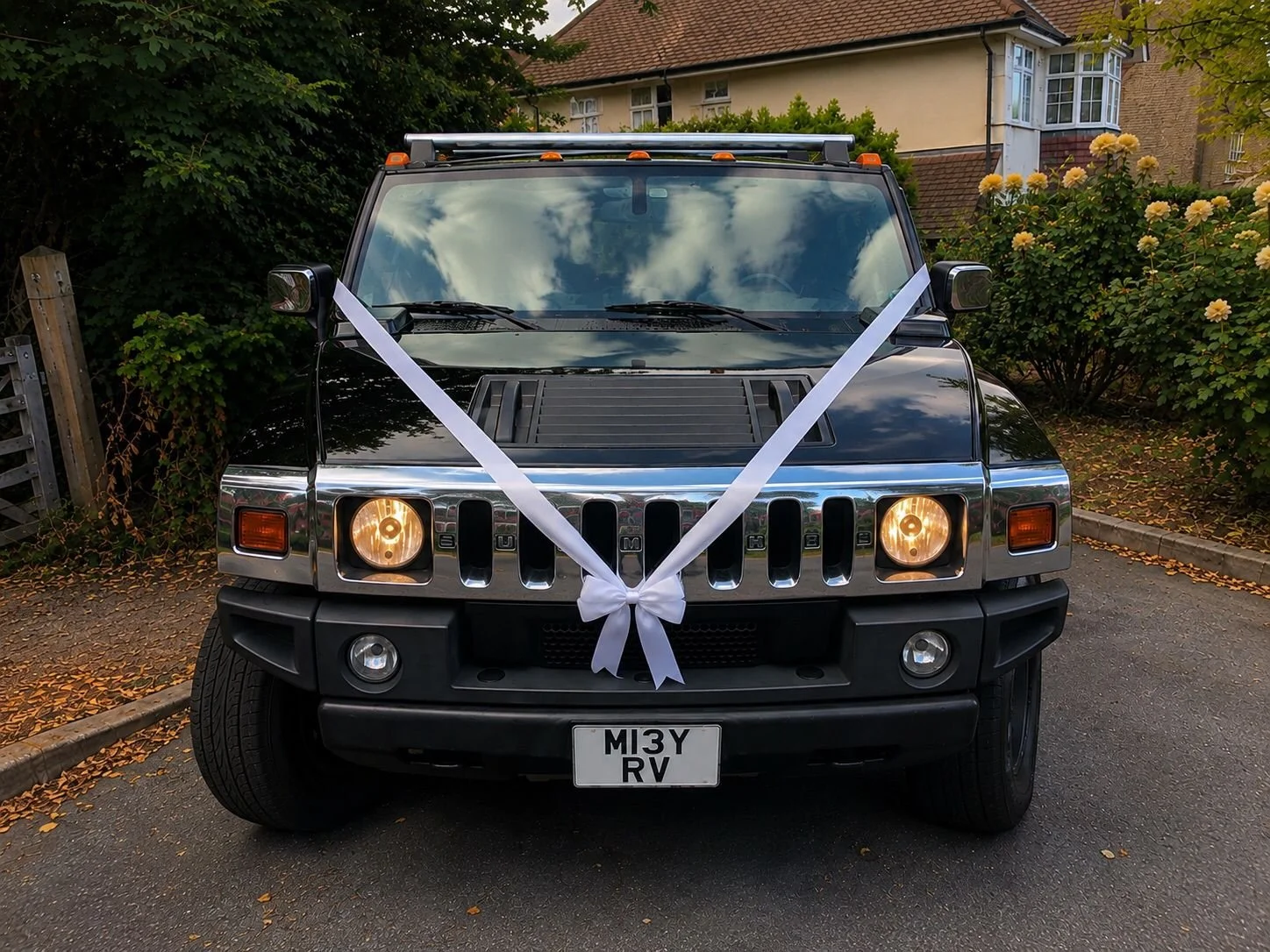 Black Jeep with a white ribbon tied on the front, indicating it is decorated for a wedding, parked on a residential street with house and flowers in the background.