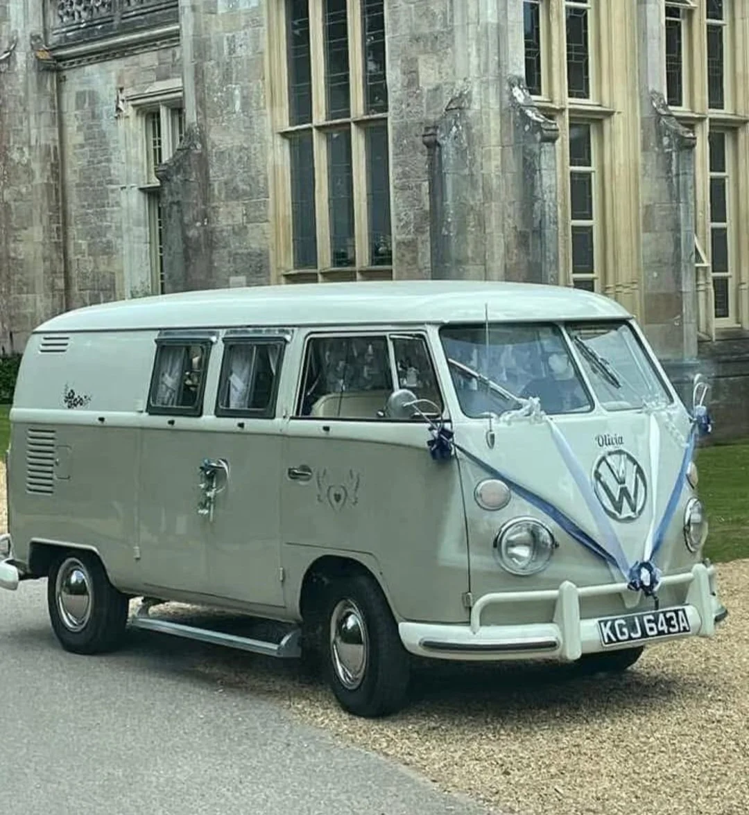 A vintage Volkswagen Type 2 van decorated for a wedding with blue ribbons and bows, parked outside a stone building with large windows.