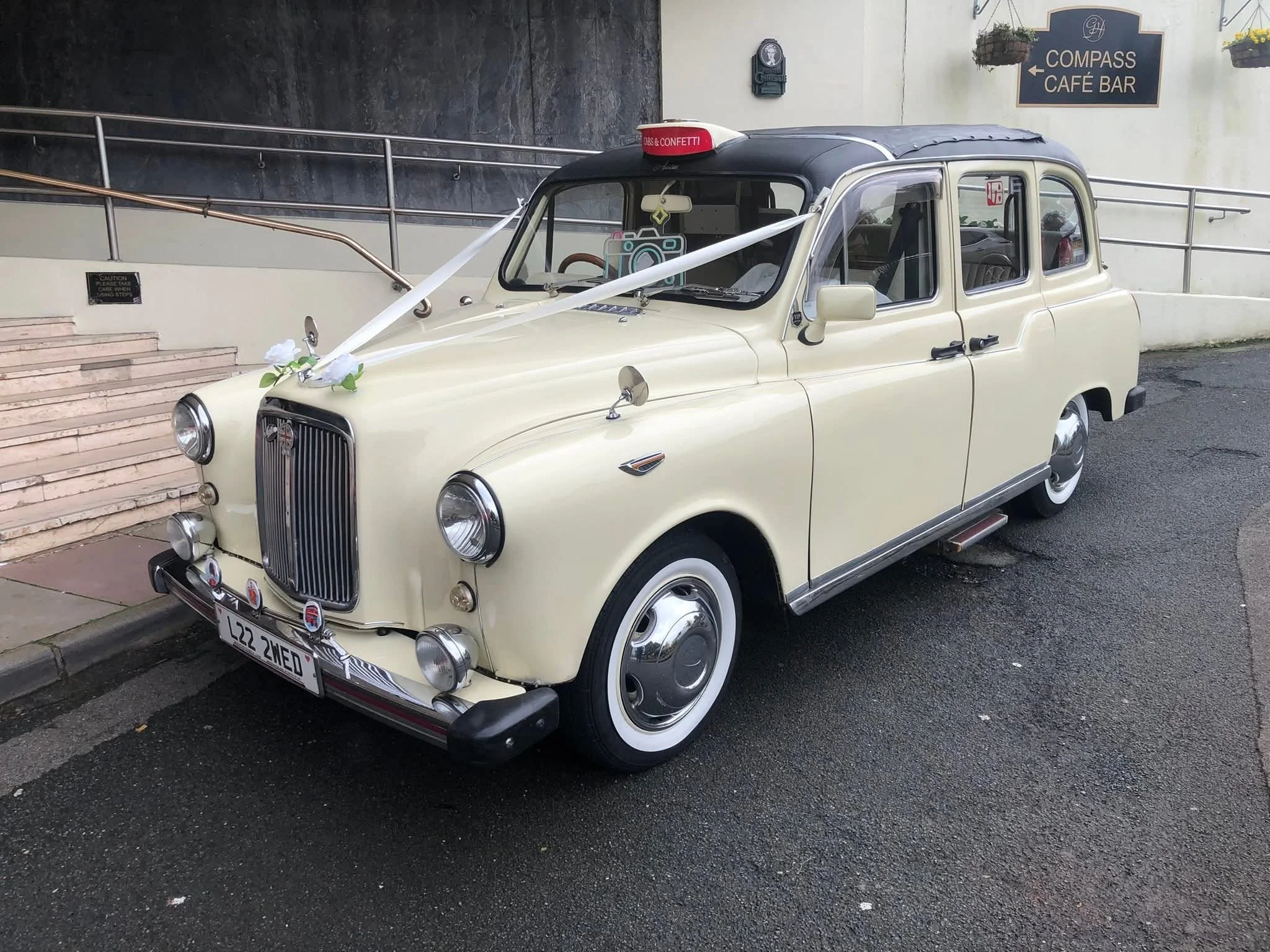 Vintage beige taxi with a wedding decoration, including white ribbons and paper flowers on the hood, parked on the street near a café bar.