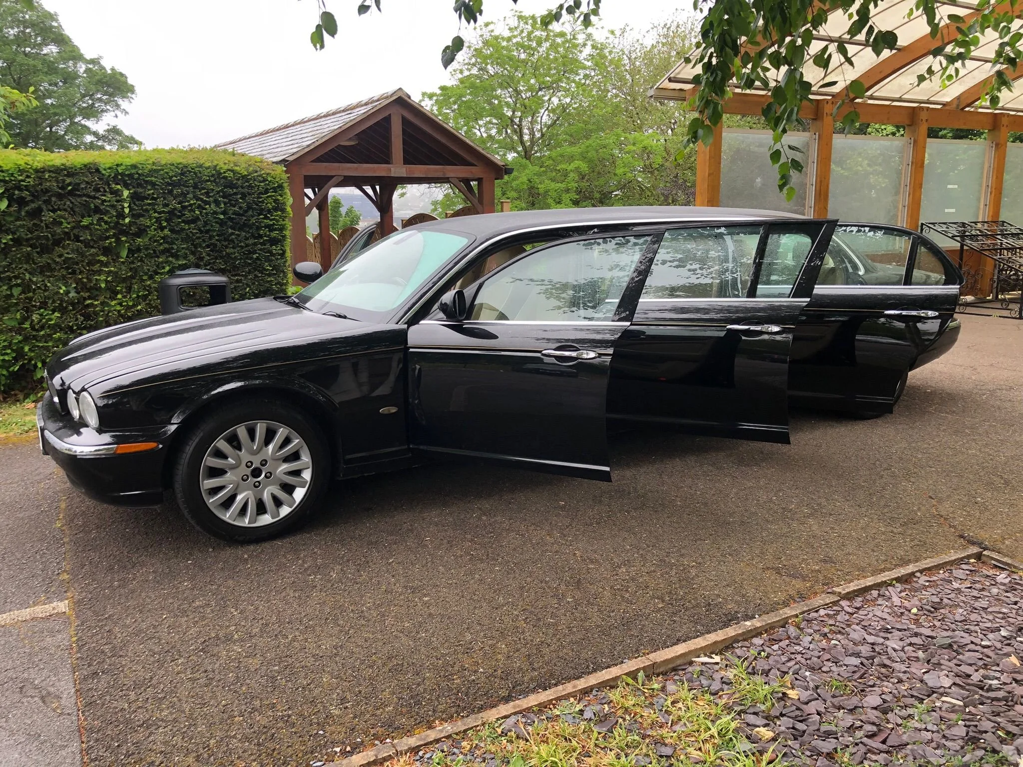Black luxury sedan car parked on a paved driveway near a wooden pergola and fence, with greenery and trees in the background.