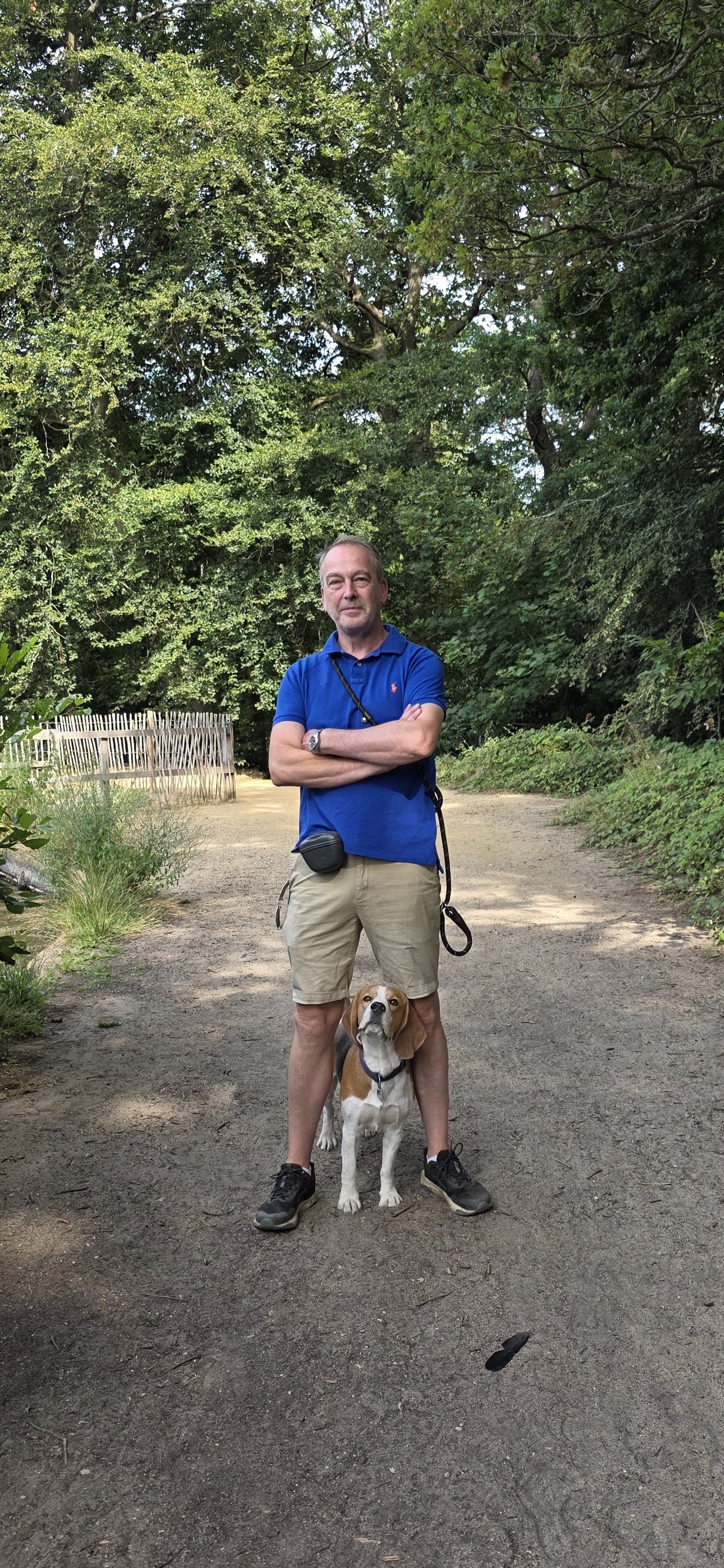 A man in a blue polo shirt and beige shorts standing on a dirt path with his arms crossed, accompanied by a beagle dog standing in front of him, set in a lush green forest.