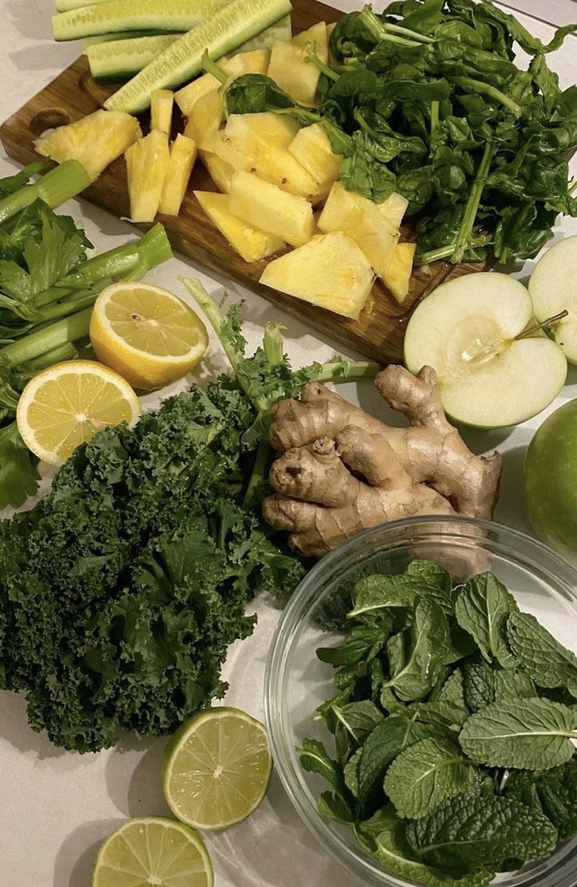 A variety of fresh fruits and vegetables, including pineapple, cucumber, green apples, ginger, lemon, kale, and mint leaves, arranged on a table.