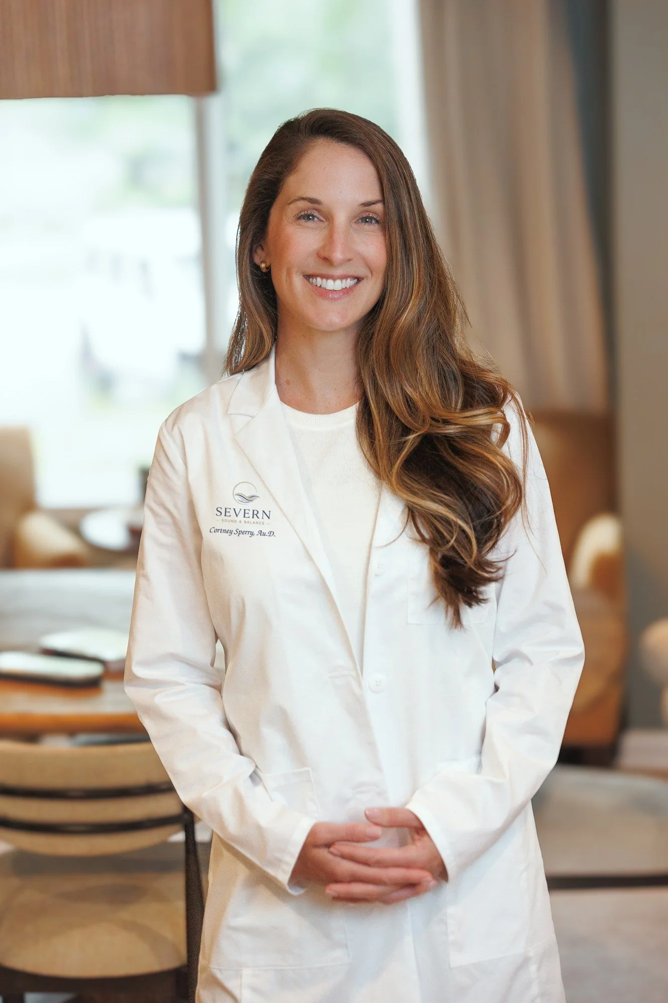 A smiling woman with long brown hair wearing a white lab coat, Dr. Sperry is a local friendly audiologist.