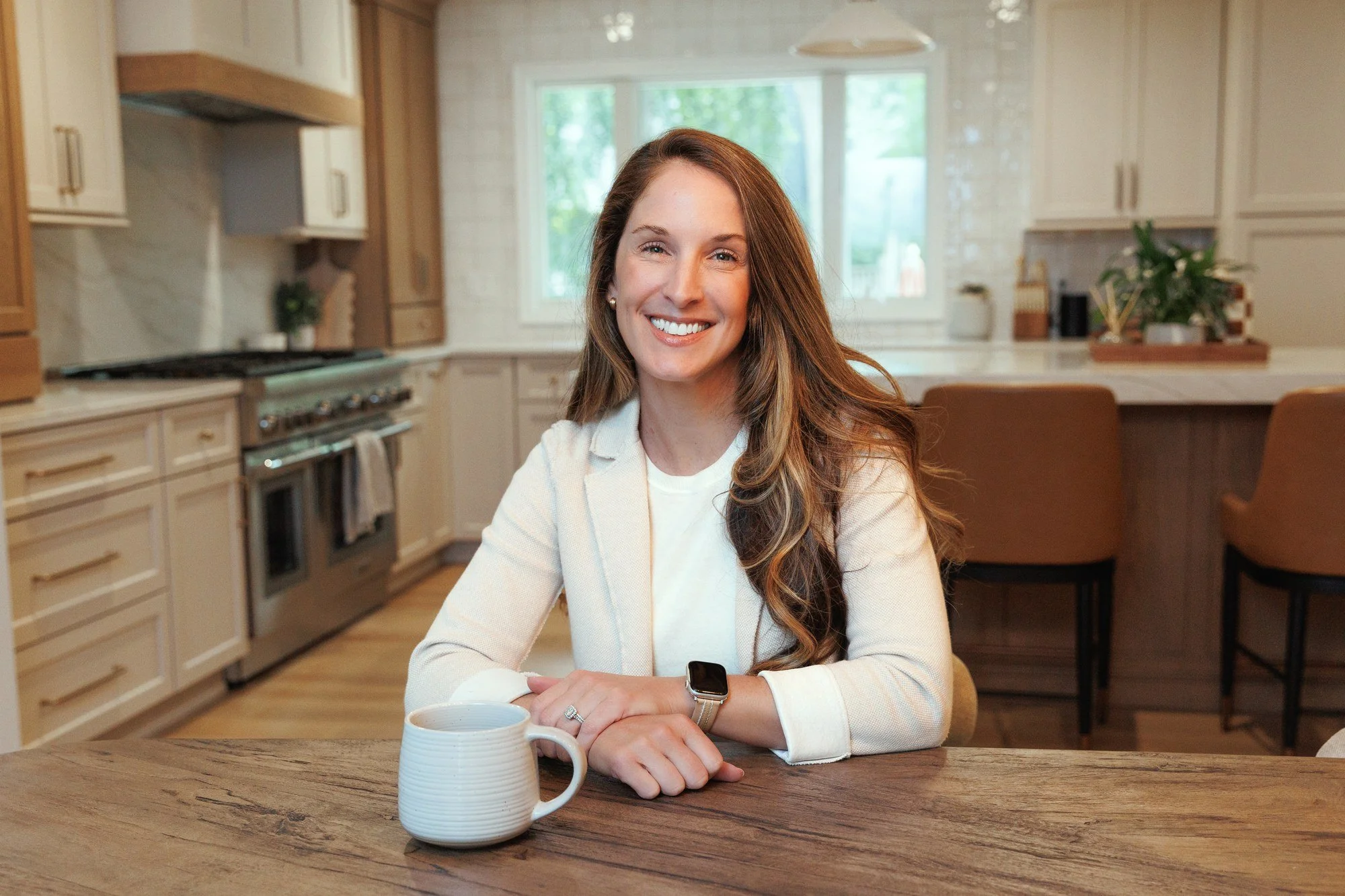 Dr. Sperry, local audiologist, sitting at a wooden table in a kitchen, smiling, with a mug and a coffee cup in front of her- at home hearing testing.