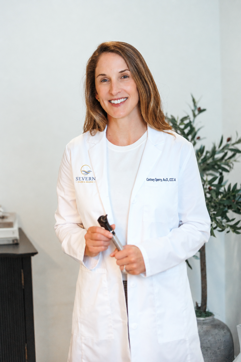 Female audiologist in a white lab coat smiling at the camera while holding an otoscope for checking individual's ears.