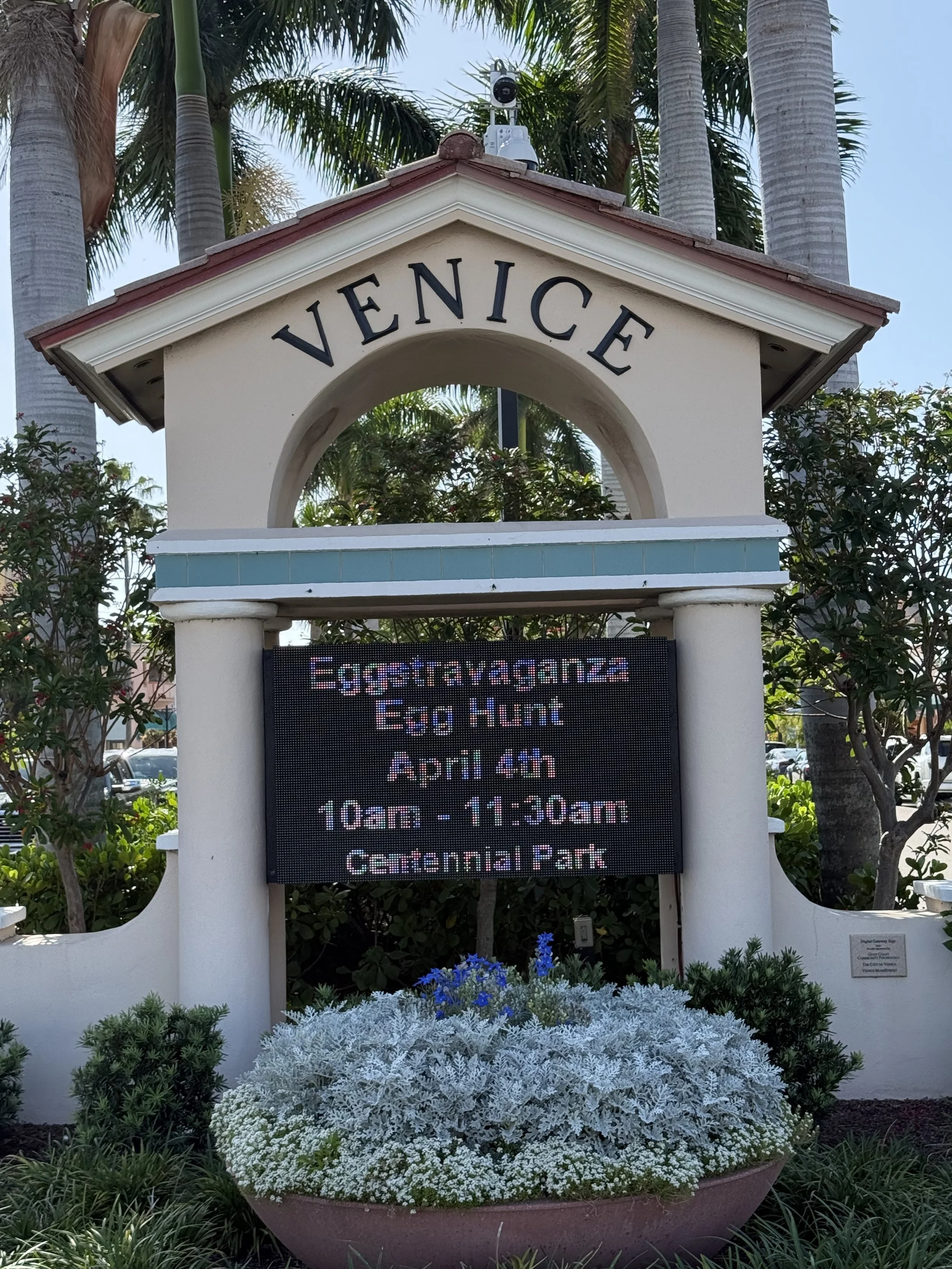 A white sign with the word 'VENEICE' at the top, situated within a small pavilion with a red roof, surrounded by green plants and palm trees. Below the sign, there is an electronic display board announcing an egg hunt event on April 4th from 10 am to 11:30 am at Centennial Park.
