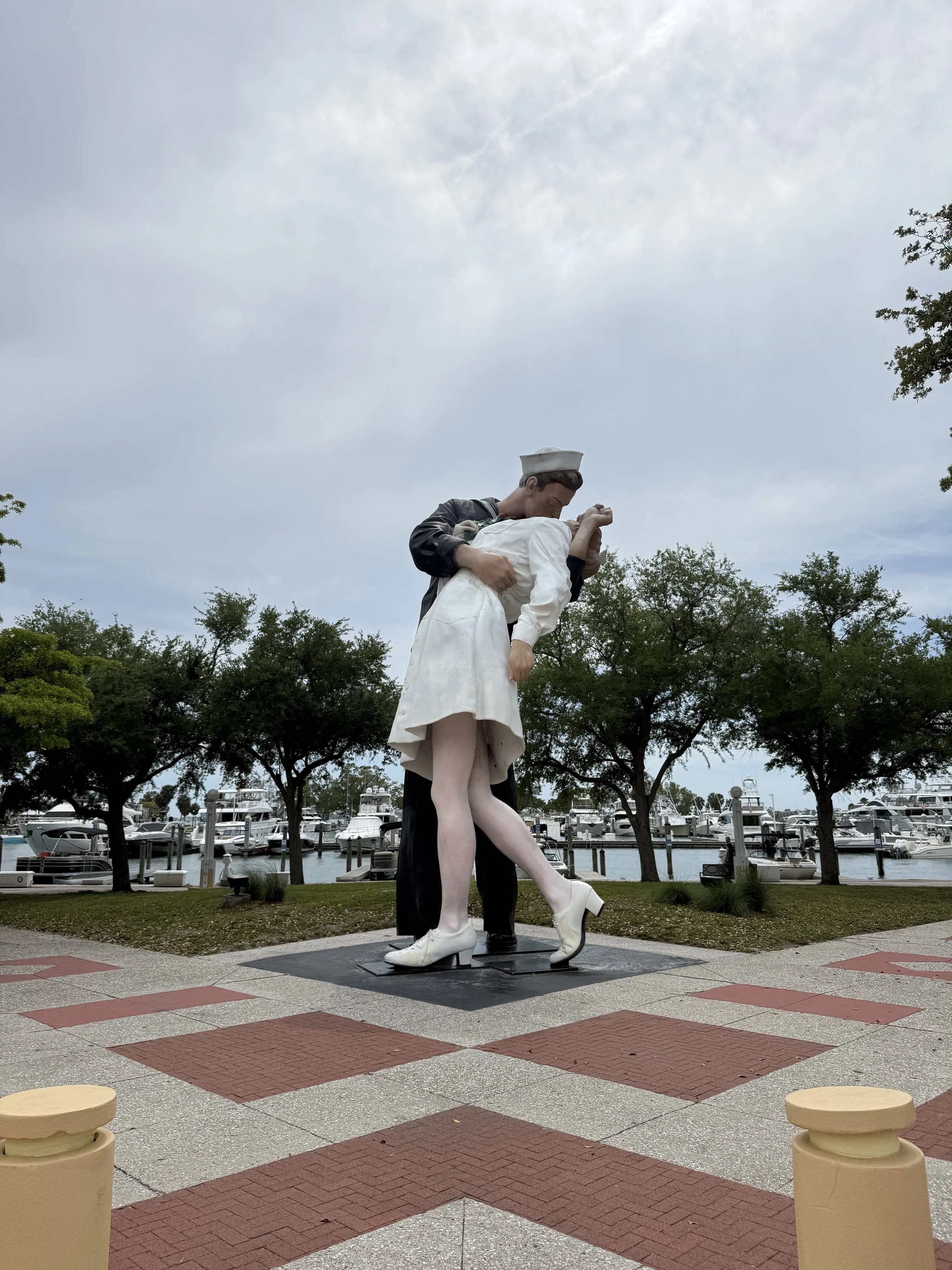 Statue of a nurse and a soldier dancing, located near a marina with boats, trees, and a cloudy sky in the background.
