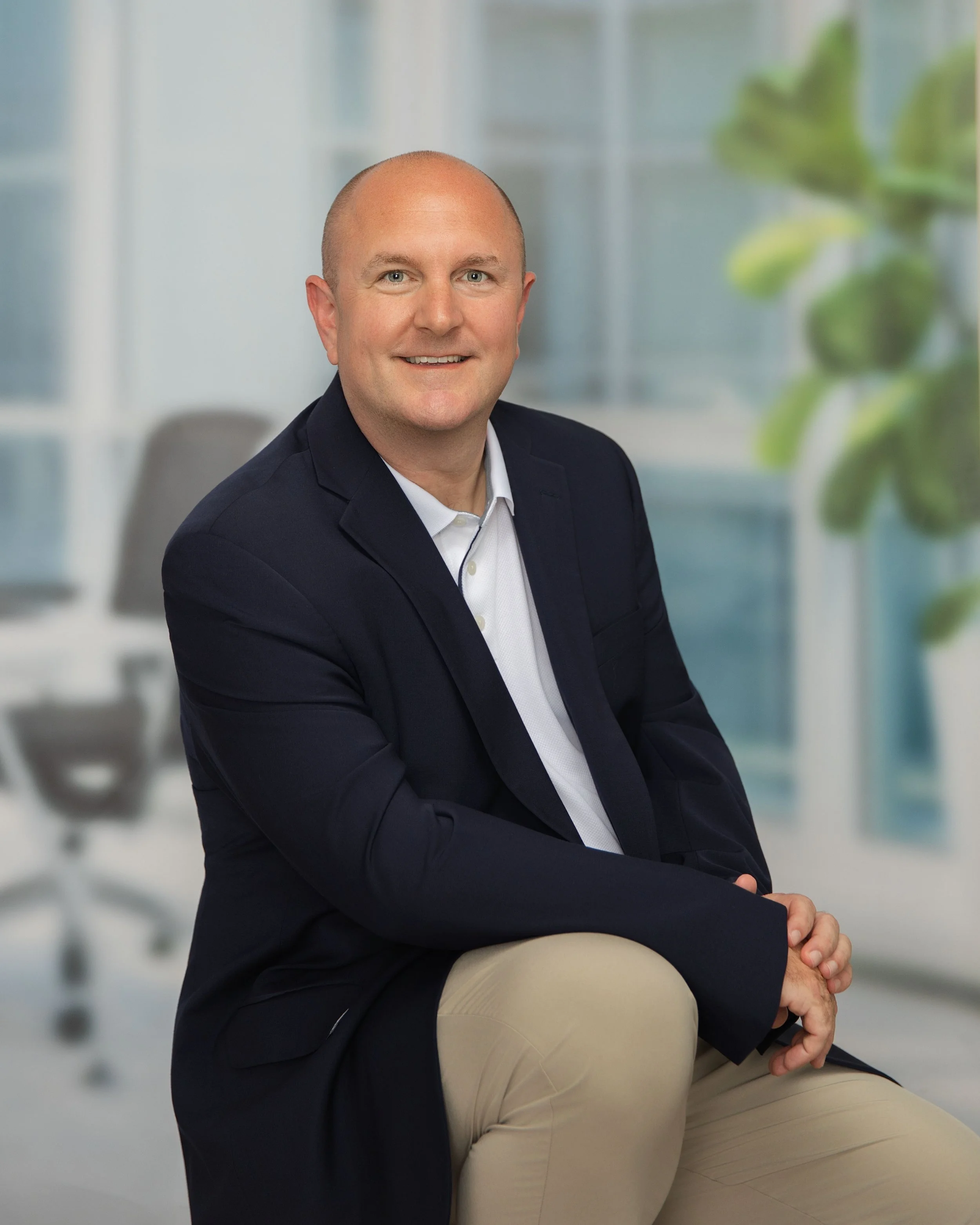 A man in a navy blazer and white shirt sitting in a modern office with blurred furniture and a green plant in the background.