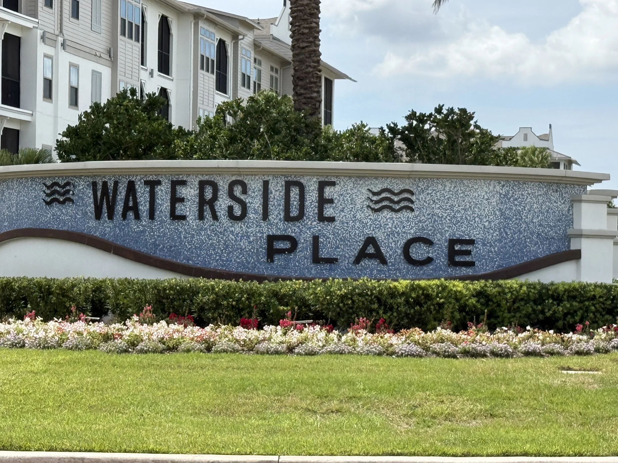 Sign reading 'WATER'S EDGE PLACE' with wavy line symbols on a landscaped area with green grass, colorful flowers, and bushes, with residential buildings and a cloudy sky in the background.