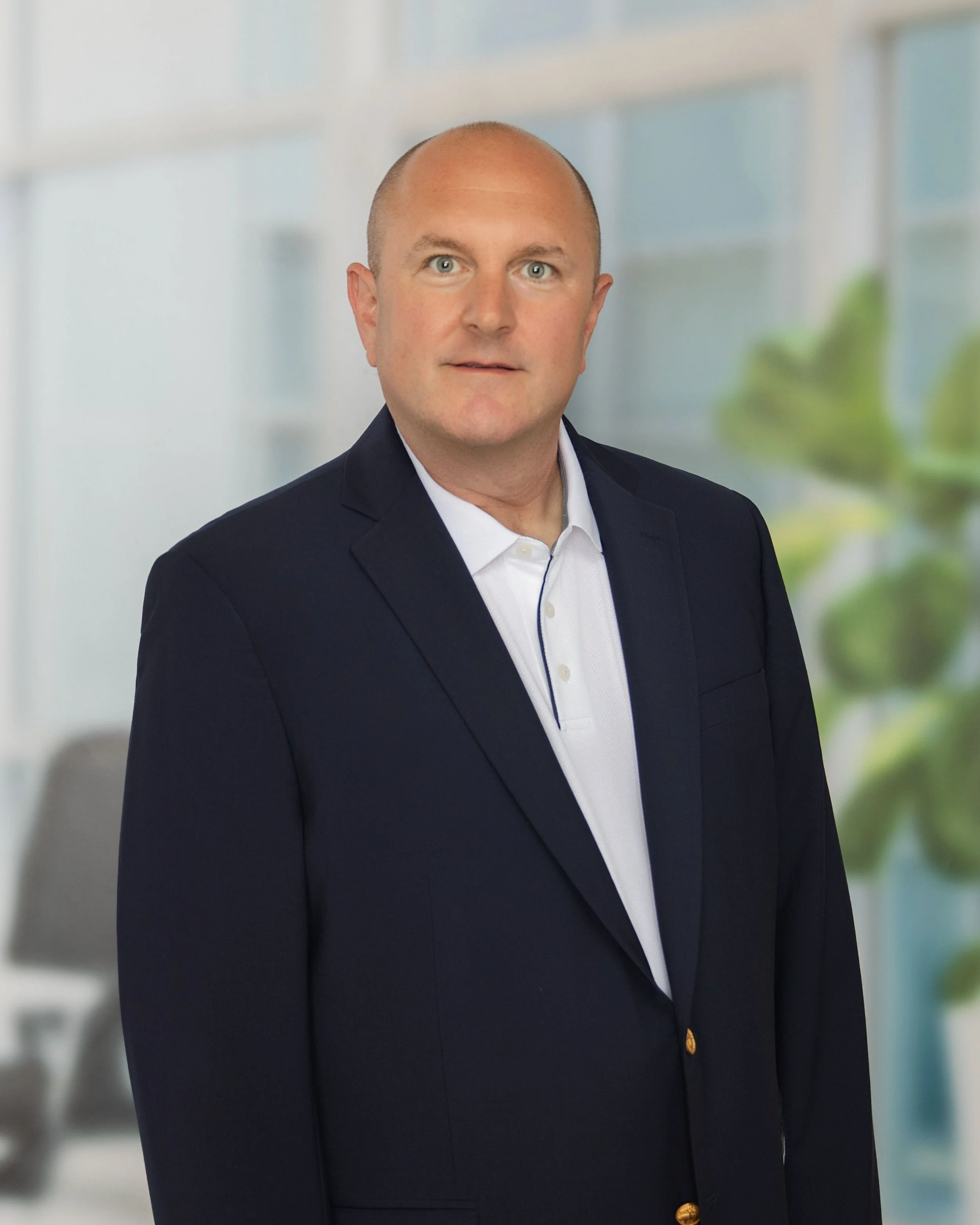 A man in a white collared shirt and dark blazer standing in an office setting with blurred windows and greenery in the background.