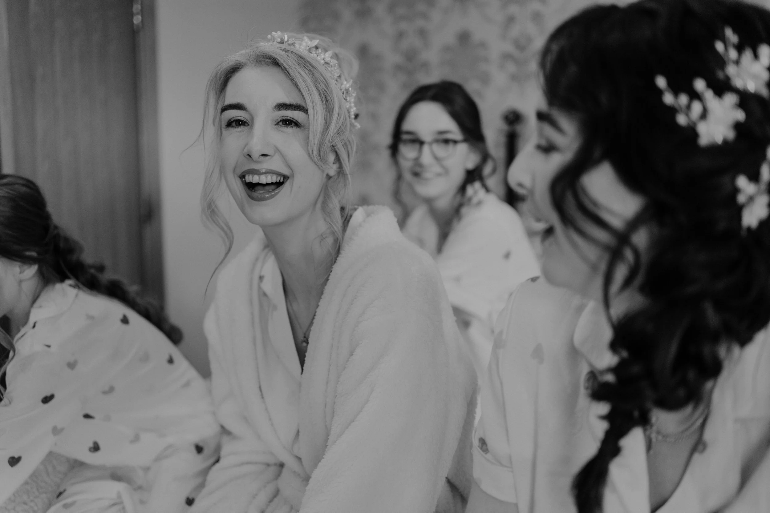 Women in robes, one with a tiara, smiling and talking, preparing for a wedding.