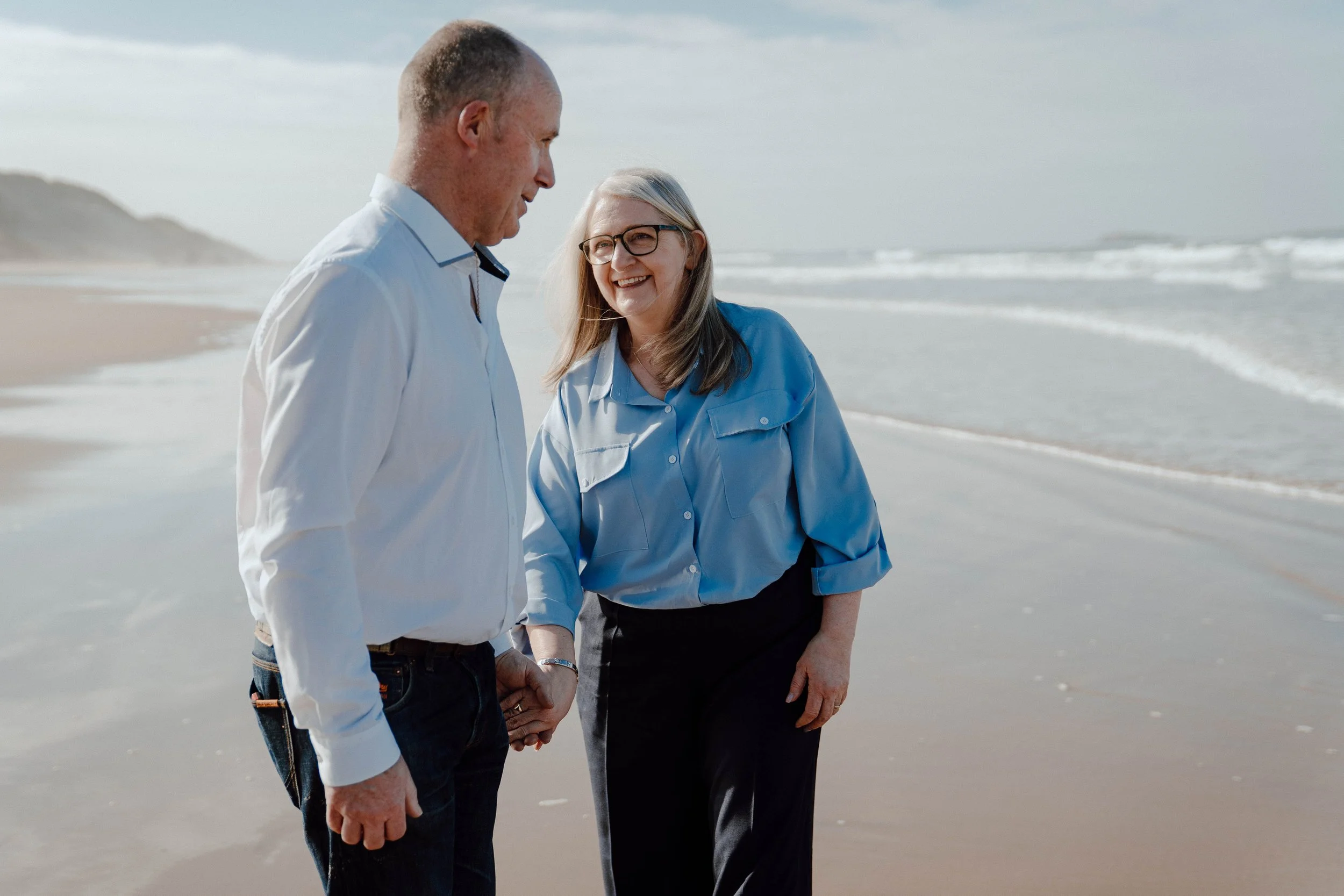 A smiling woman with gray hair and glasses holding hands with a man on a beach with ocean waves in the background.