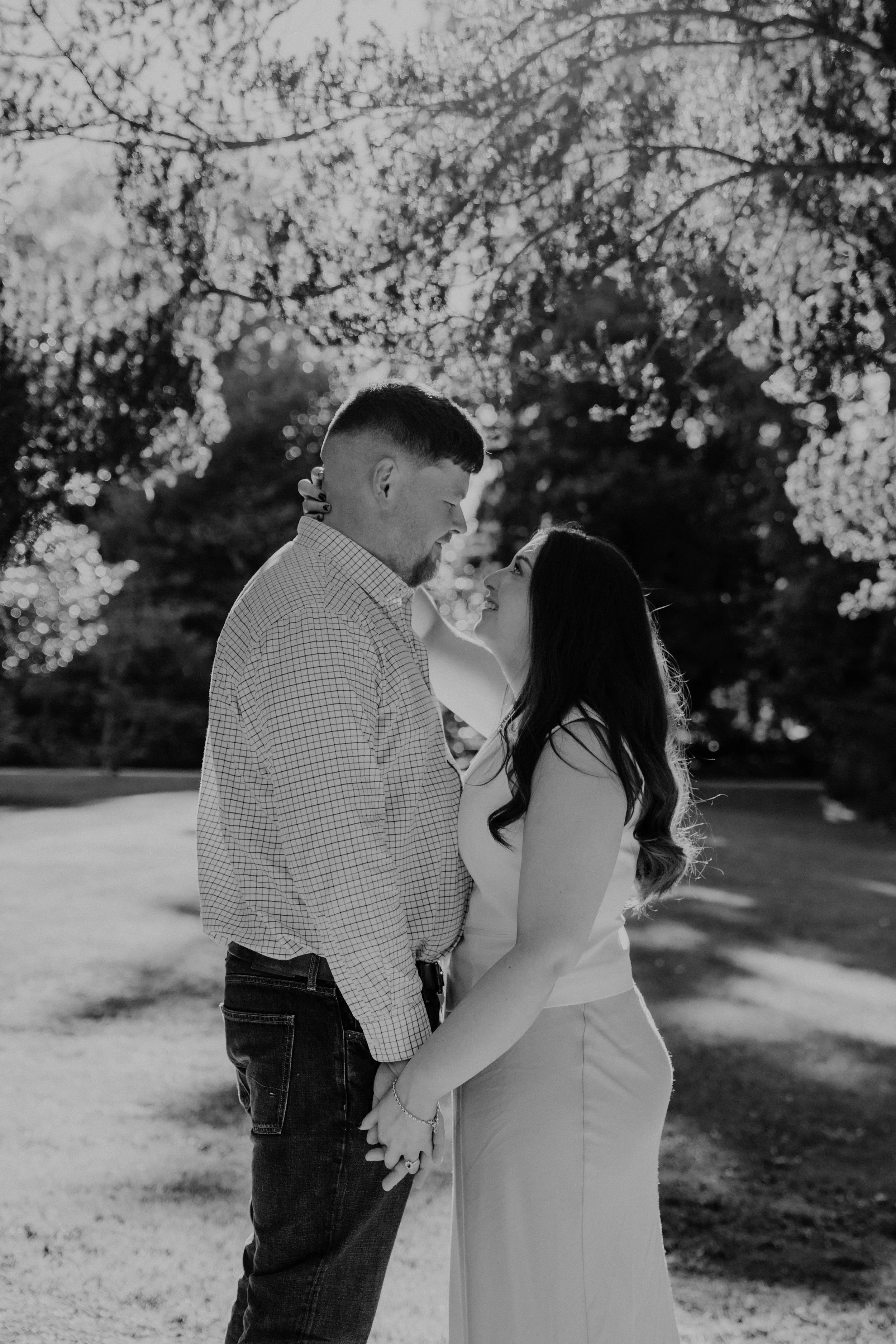 A couple holding hands and gazing into each other's eyes outdoors on a sunny day, with trees and foliage in the background, captured in black and white.