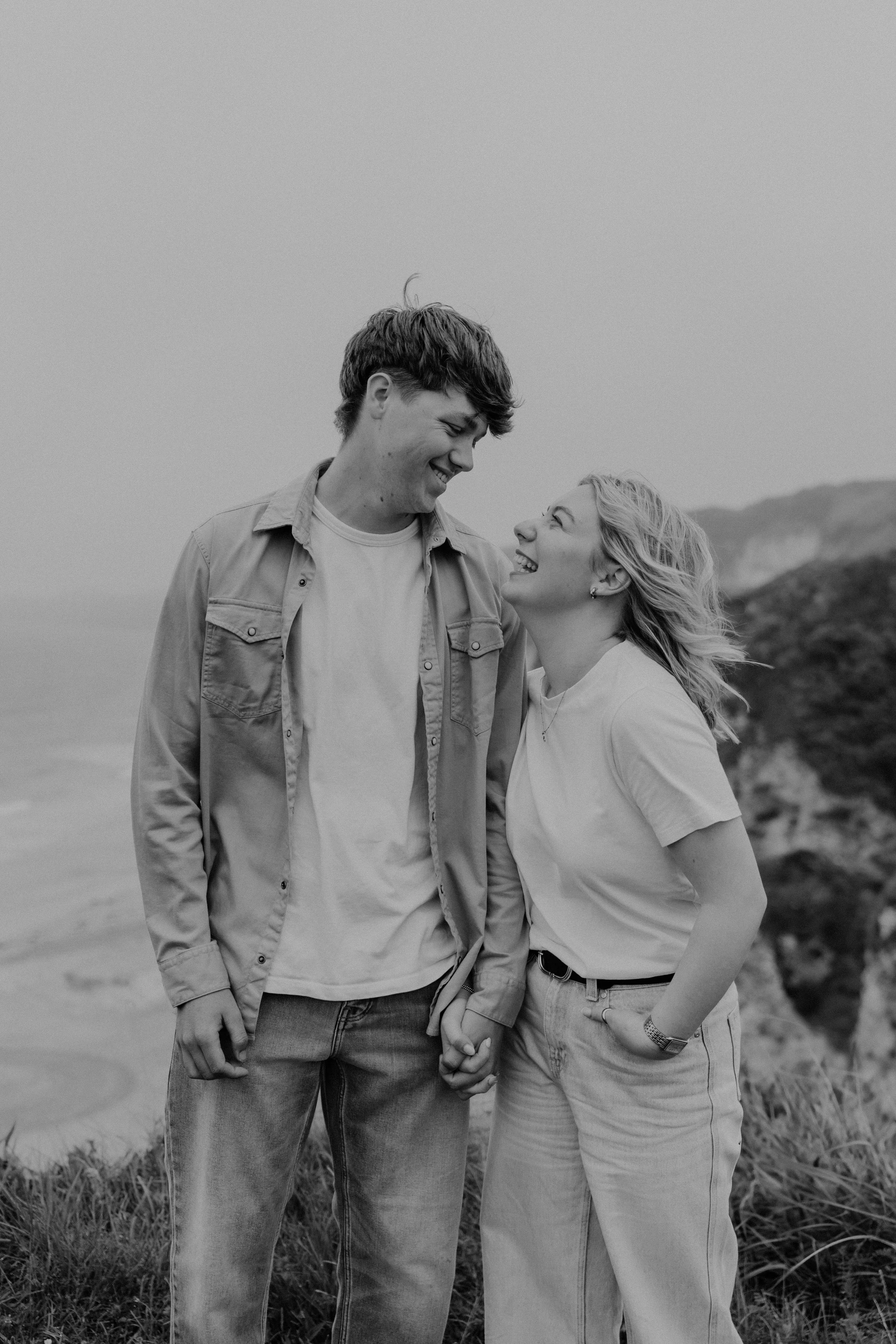 A smiling couple holding hands and facing each other on a grassy area near the beach, with cliffs and ocean in the background, in black and white.