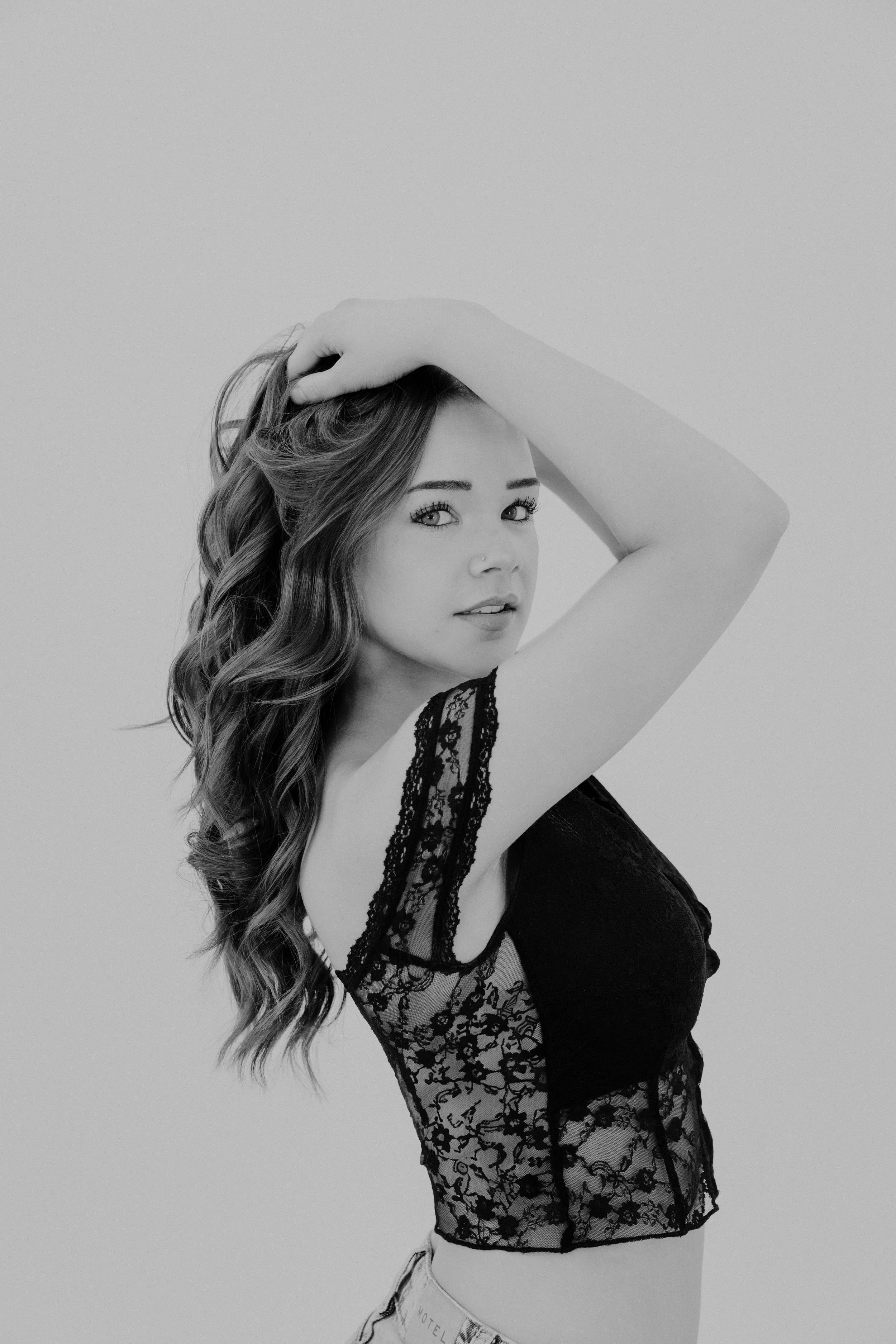 A young woman with wavy hair poses with one hand on her head in front of a plain background, wearing a lace top.
