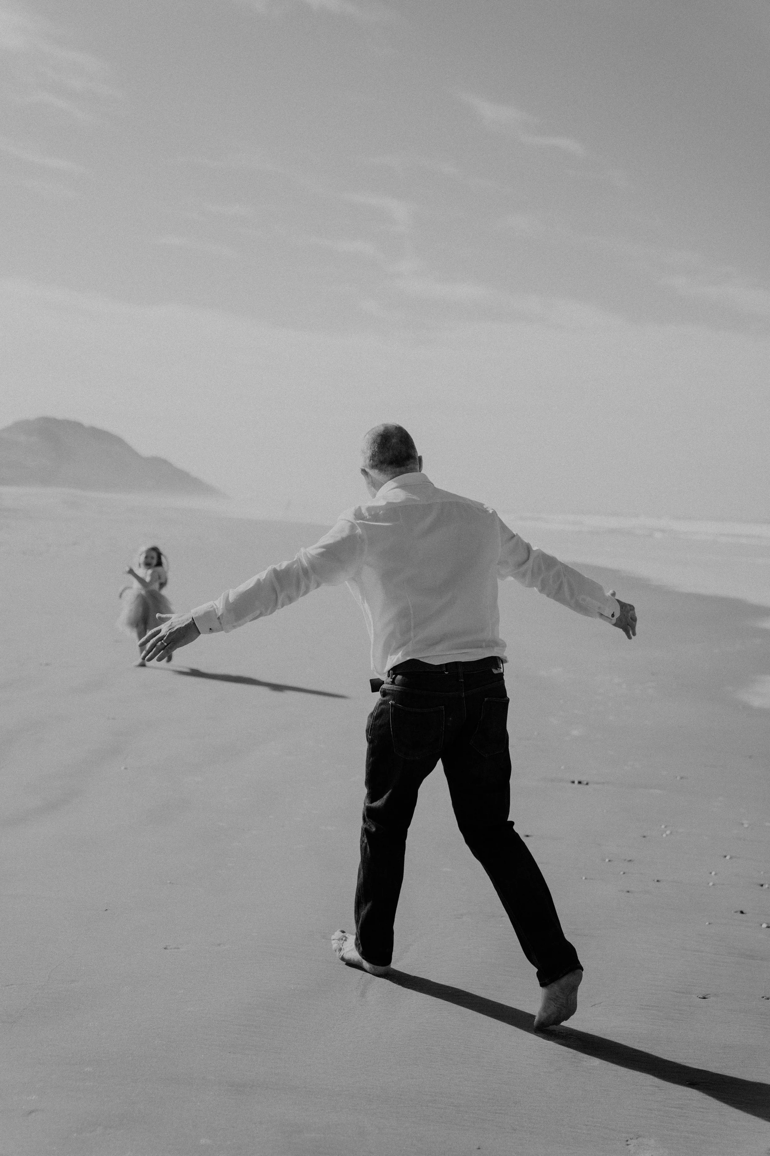 A man holding his arms out walking on the beach towards a girl in the distance, with a mountain in the background, under a sky with clouds.