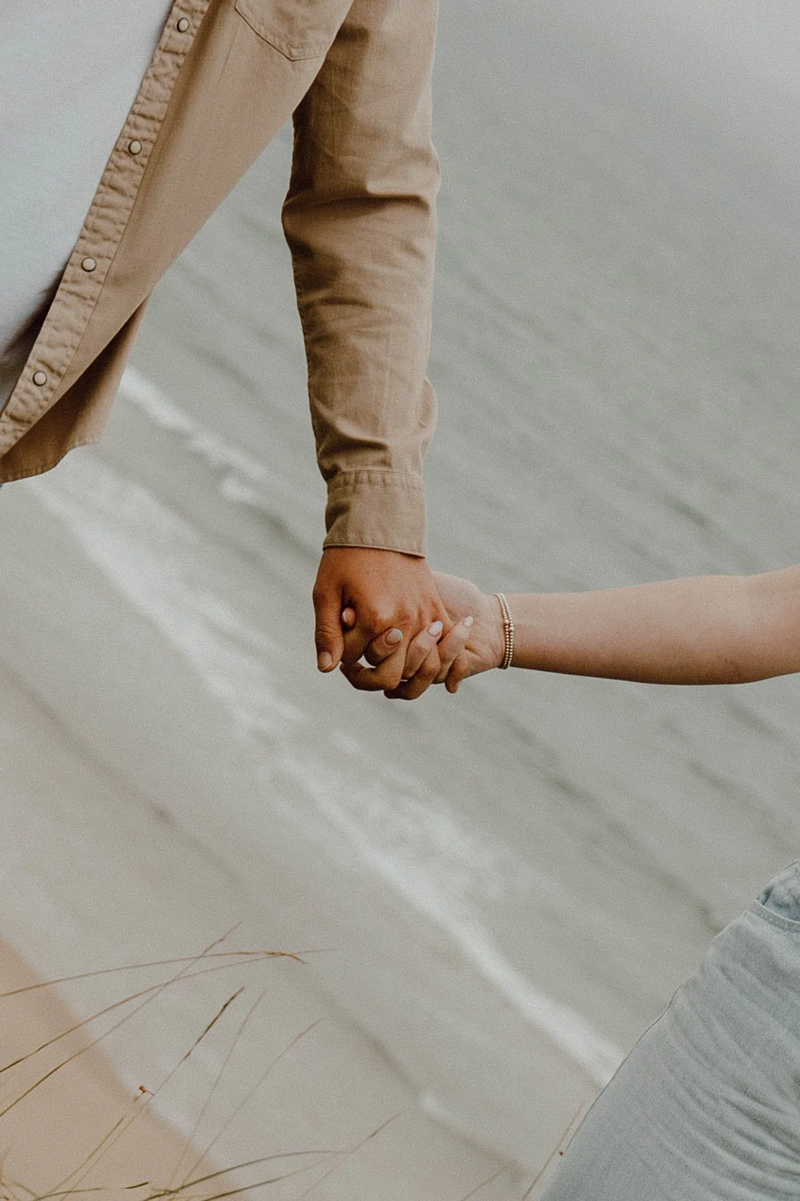 Close-up of two people holding hands, one wearing a tan long-sleeve shirt and the other wearing a bracelet, on a beach with sand and ocean waves in the background.