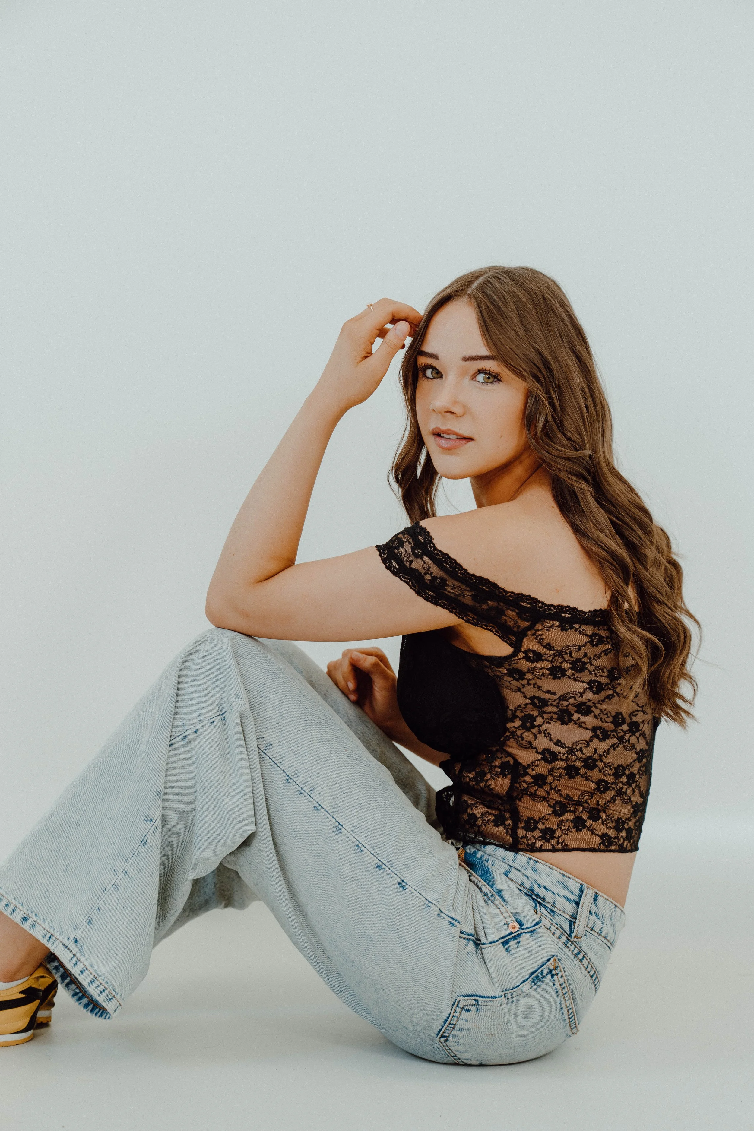 Young woman with long wavy brown hair wearing a sheer black lace top and light blue jeans, sitting with one knee bent and hand touching her hair, against a plain white background.