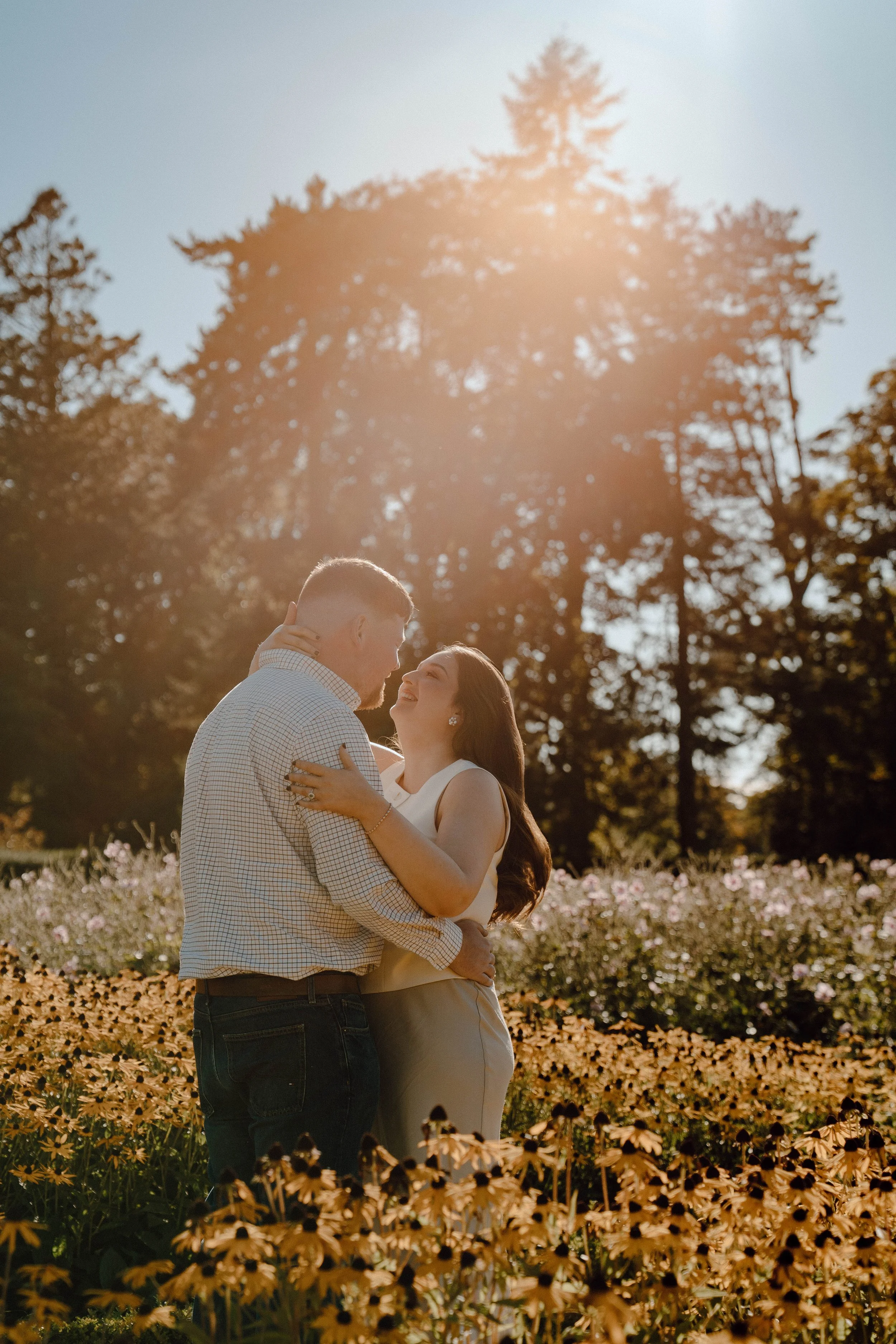 A couple embracing in a field of flowers during sunset, with trees in the background.