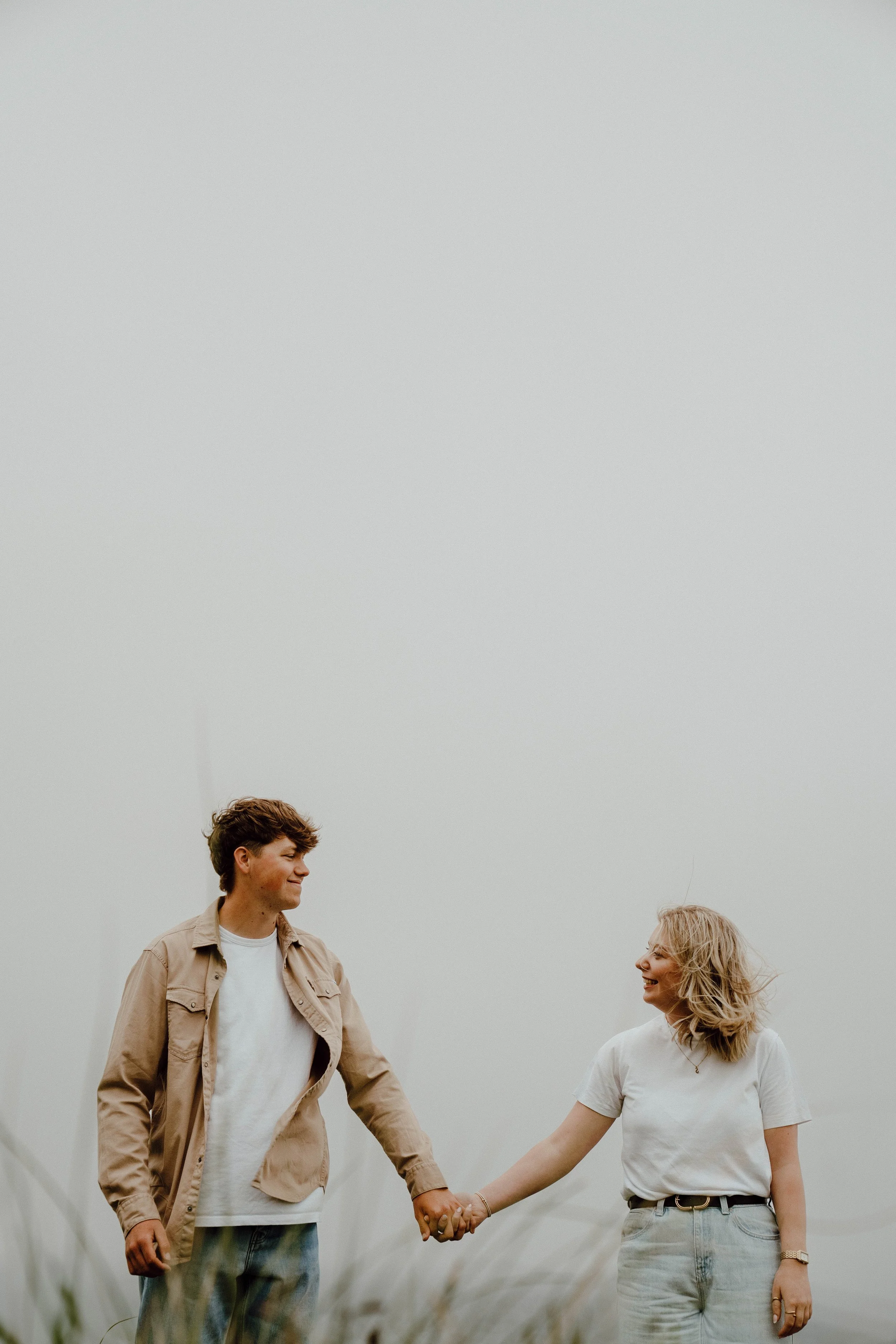 A young man and woman holding hands and smiling at each other outdoors, with foggy, overcast sky and blurred grass in the foreground.