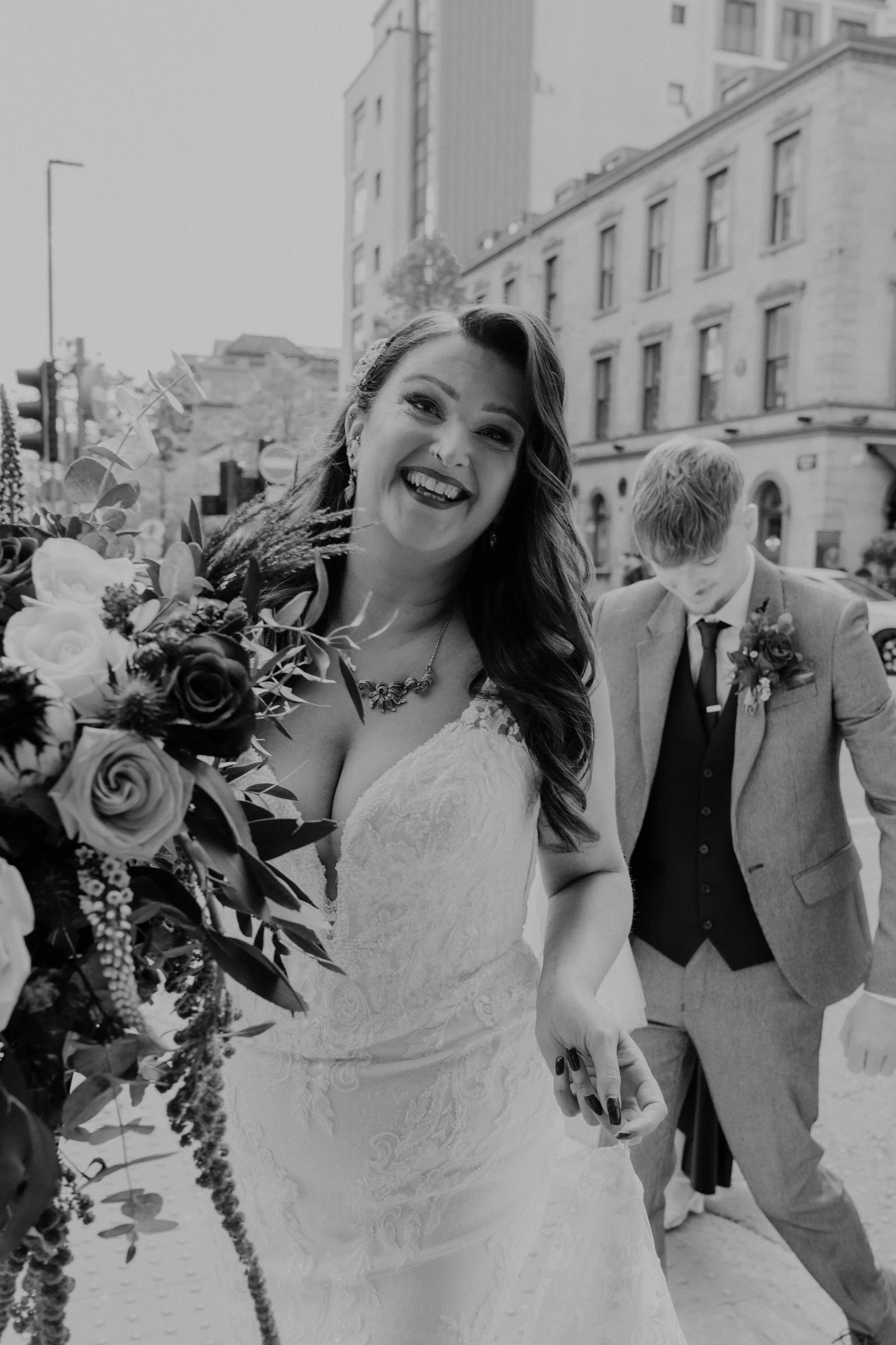 A smiling woman in a wedding dress holding a bouquet, with a man in a gray suit and young boy in the background, outside in an urban setting.