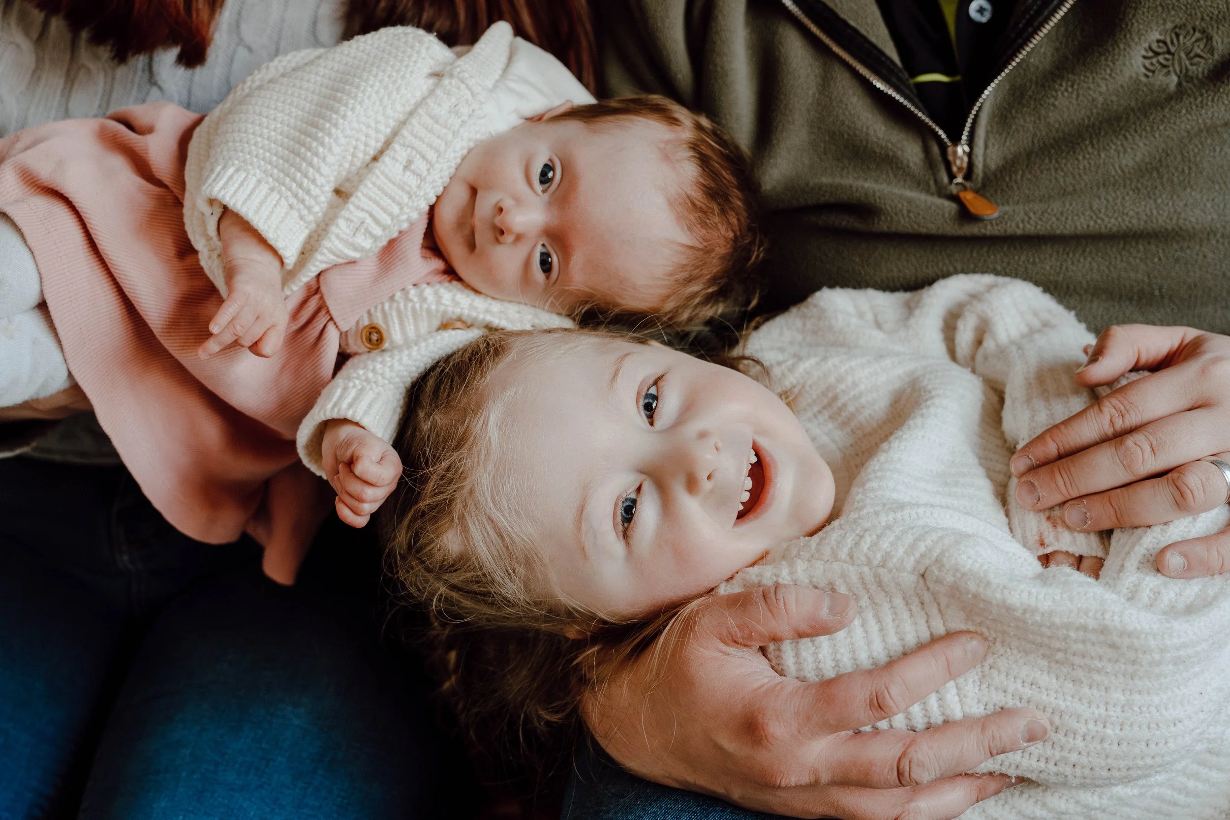 Two young children, a girl with blonde hair and an older girl with reddish-brown hair, lying down together with adults, all smiling and looking up at the camera.