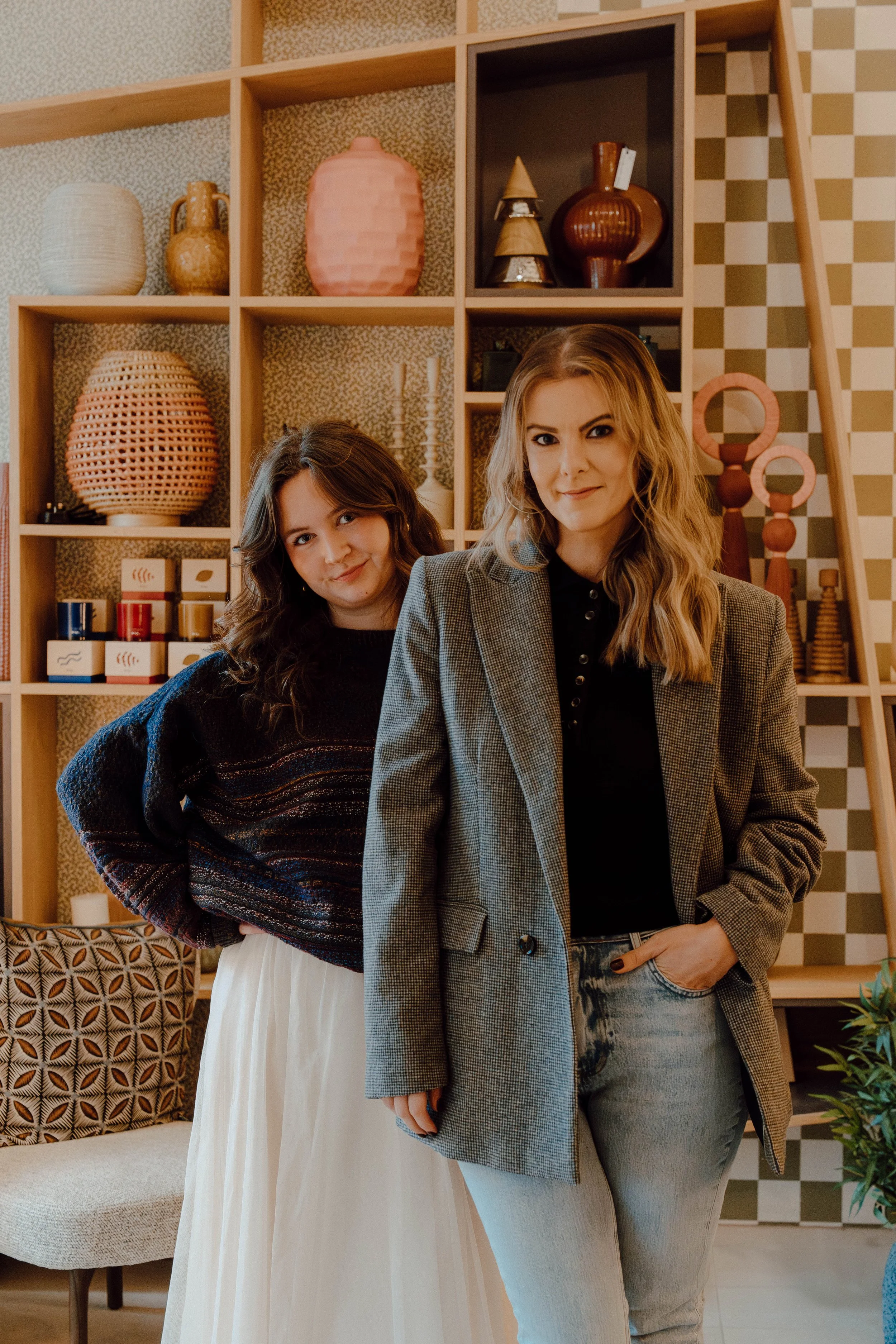 Two women standing in front of a wooden shelf filled with decorative vases and objects, smiling at the camera.