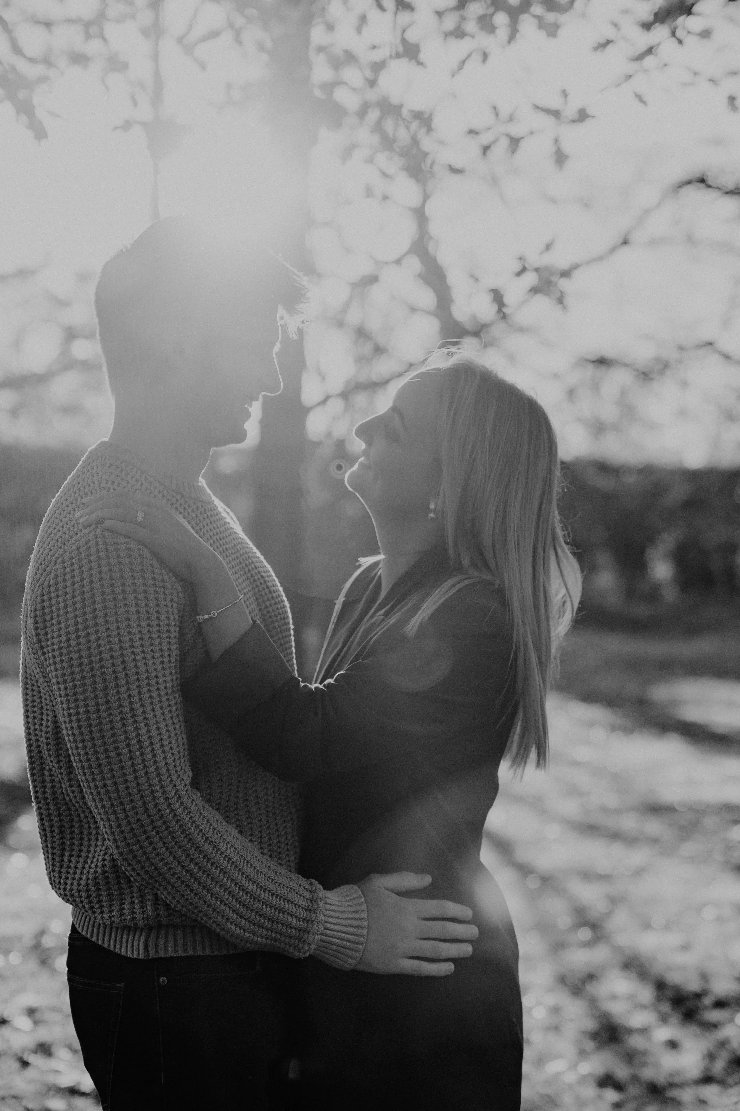 A black and white photo of a couple standing close outdoors with trees in the background. They are smiling and gazing at each other, embracing with the sun shining behind them.