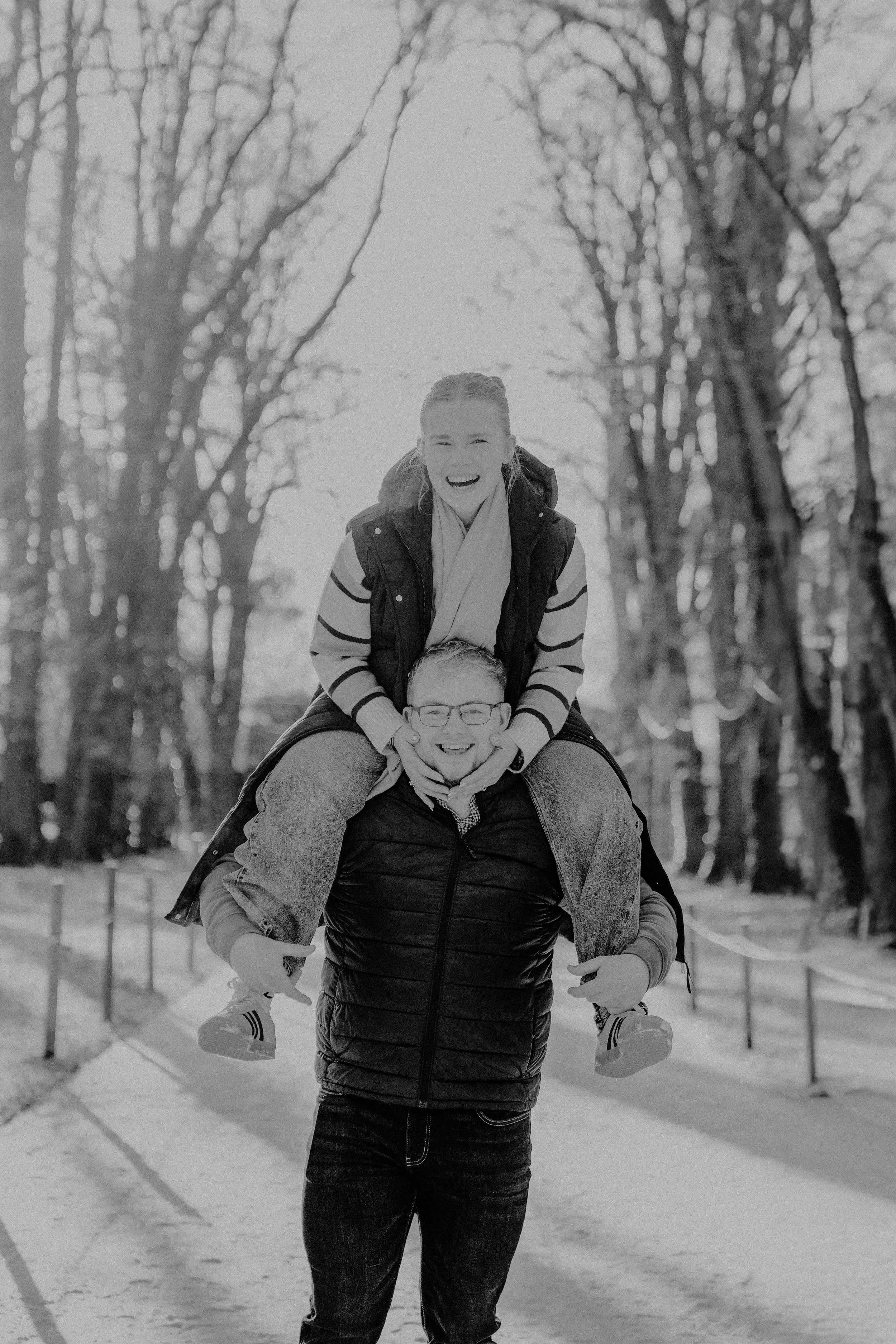 A man carrying a woman on his shoulders in a park with leafless trees, both smiling.