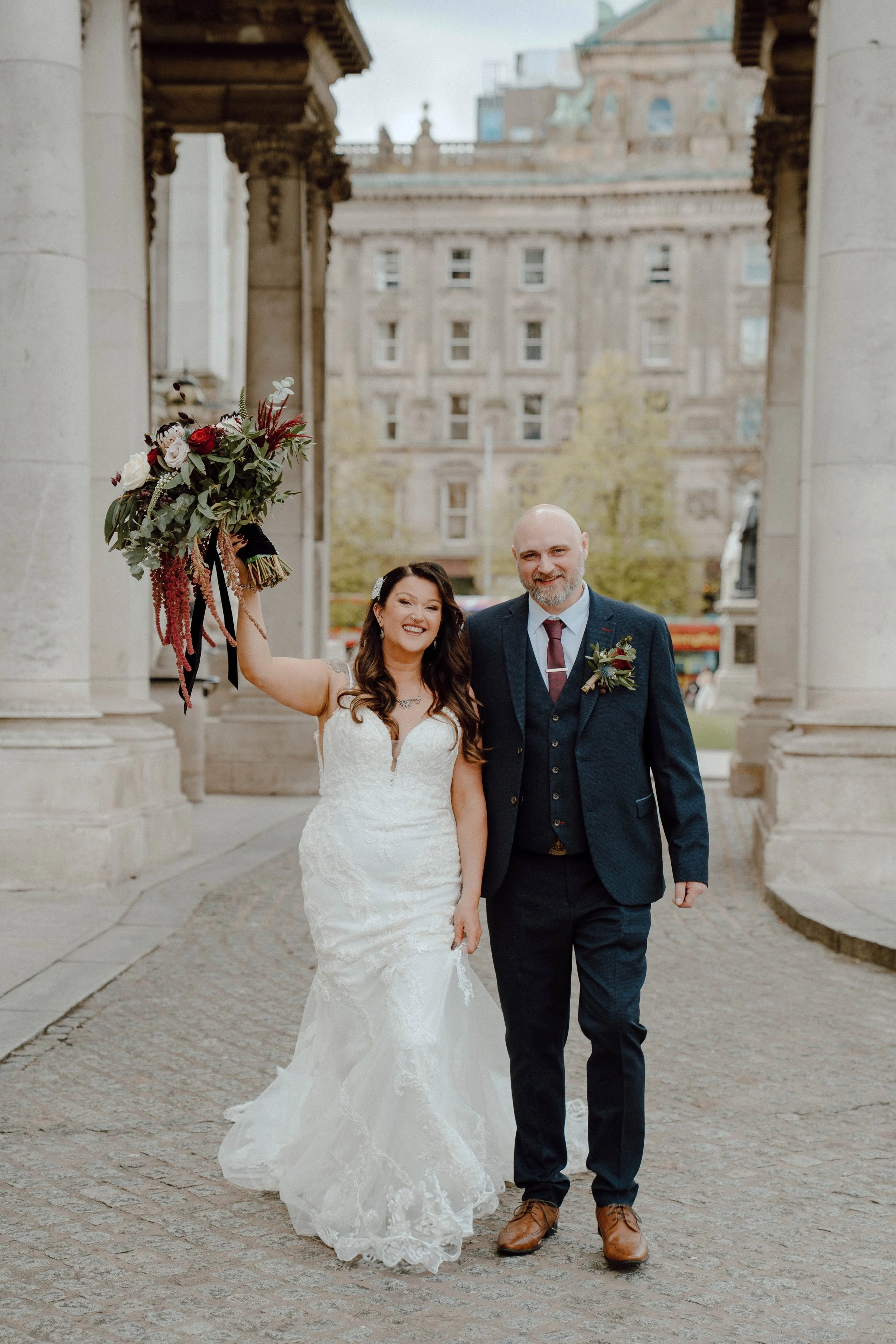 Bride and groom celebrating after their wedding, standing under a stone archway, with the bride holding a large floral bouquet and smiling.