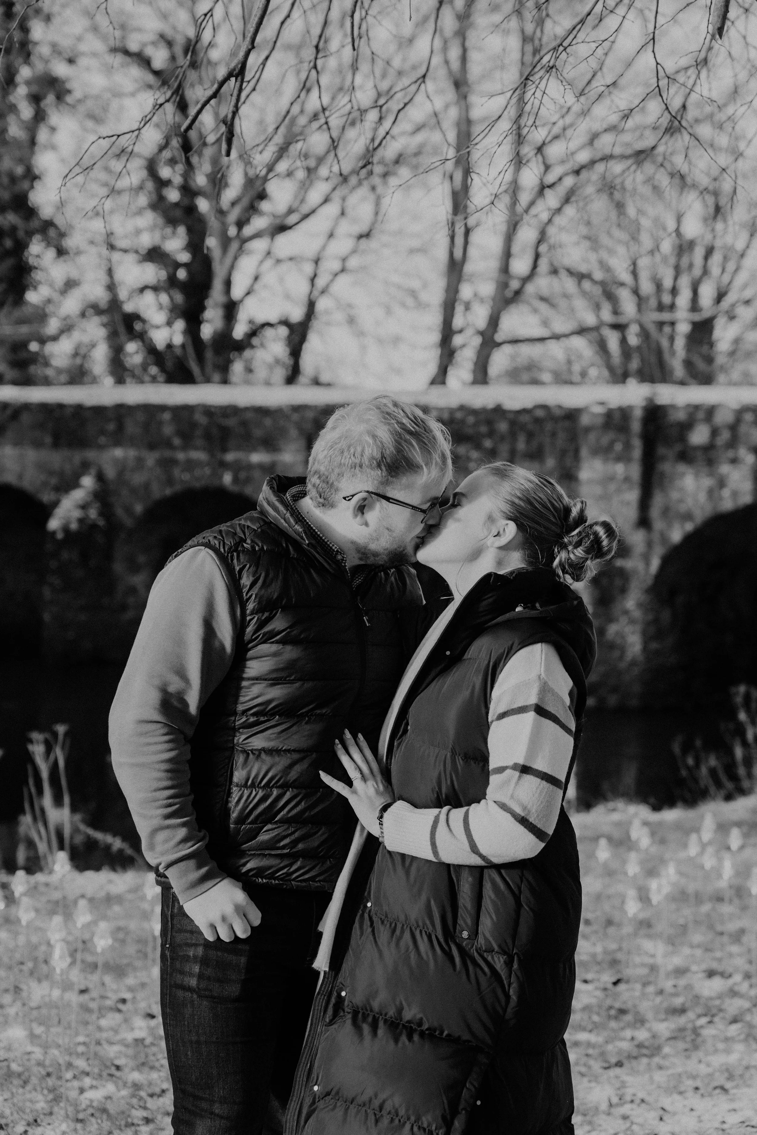 A black and white photo of a couple kissing outdoors in front of a stone bridge and leafless trees.