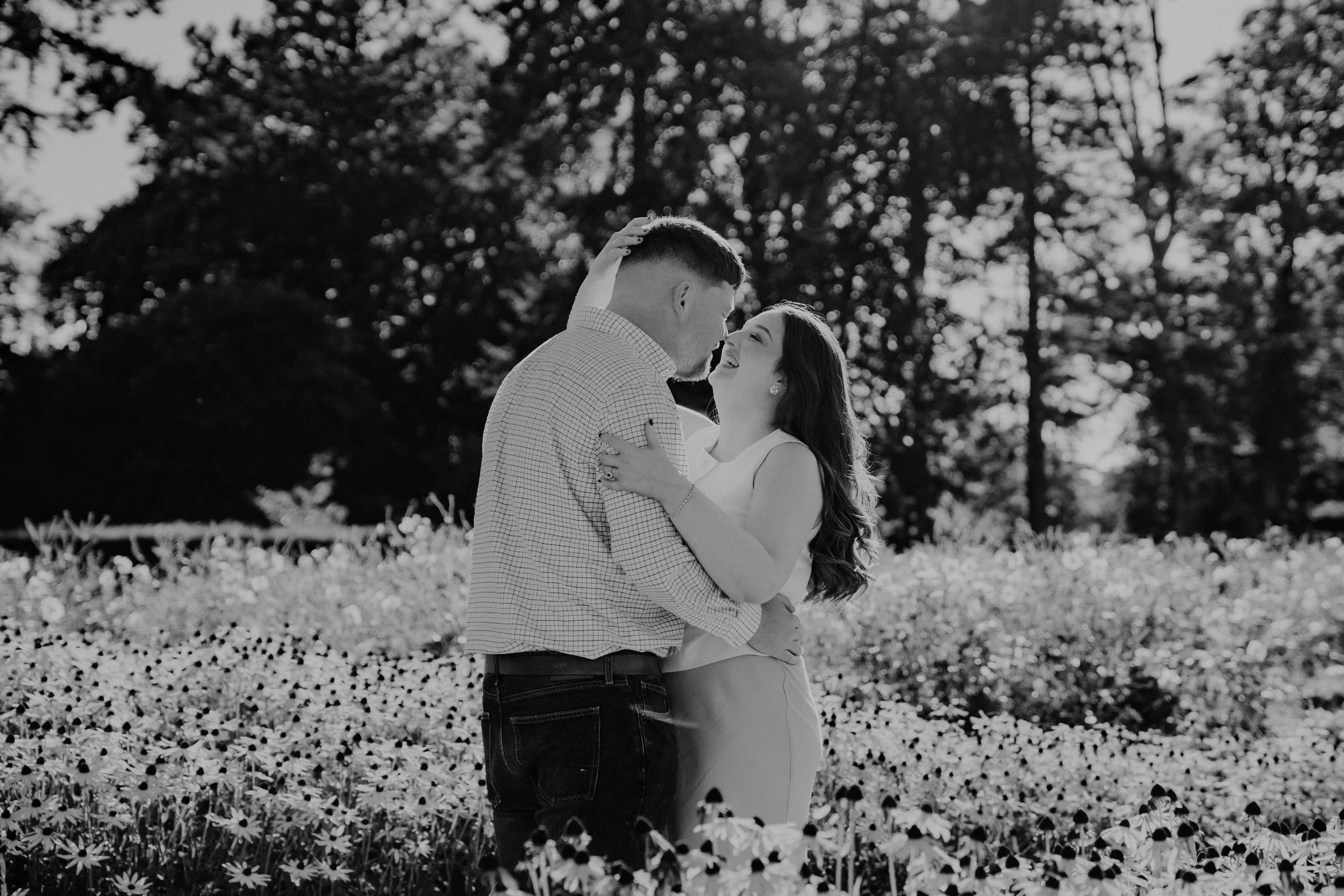 A couple sharing a romantic moment outdoors in a field of flowers, embracing and smiling at each other, with trees in the background.