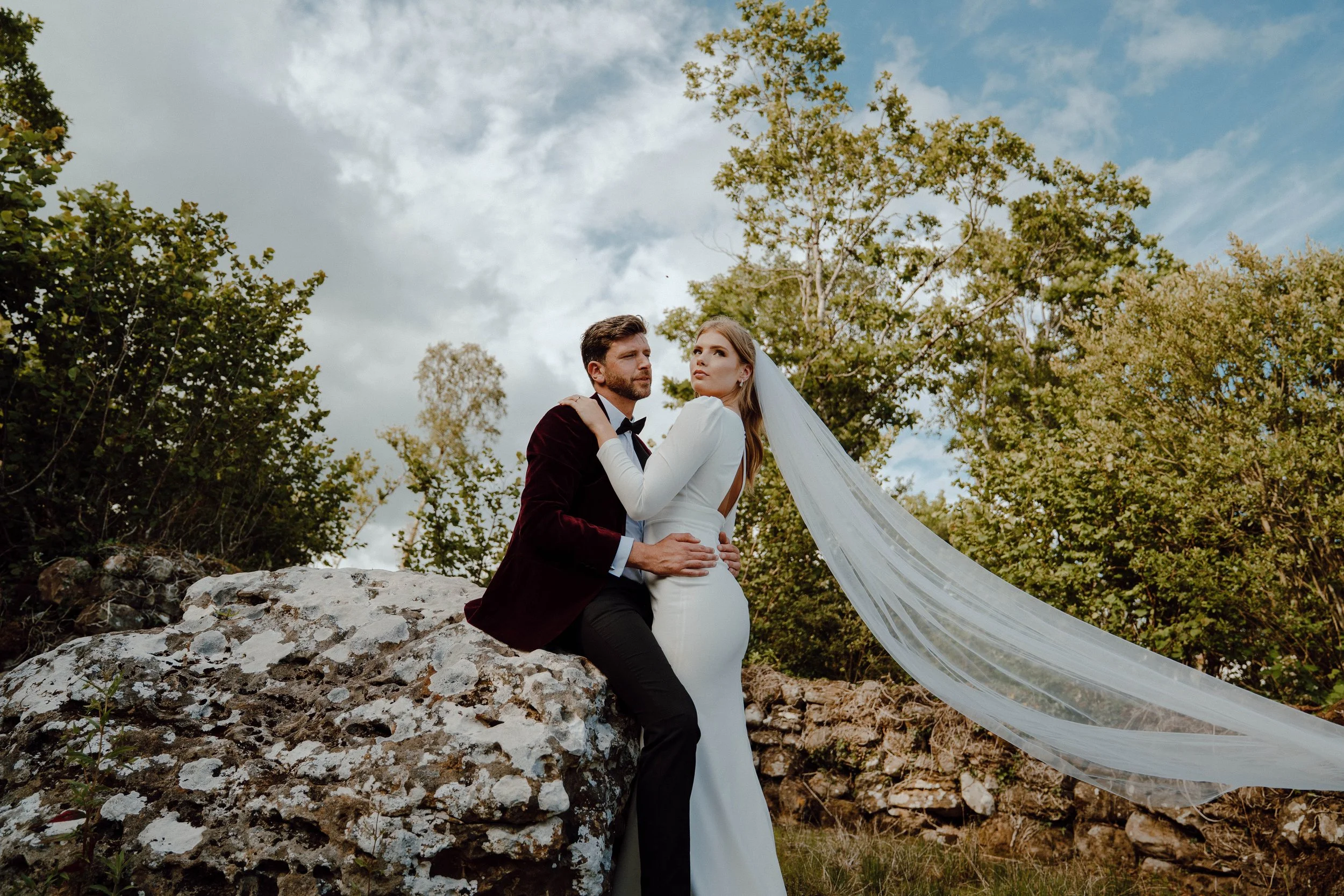 A bride and groom sitting on a rock outdoors, with trees and a cloudy sky in the background, during their wedding photoshoot.