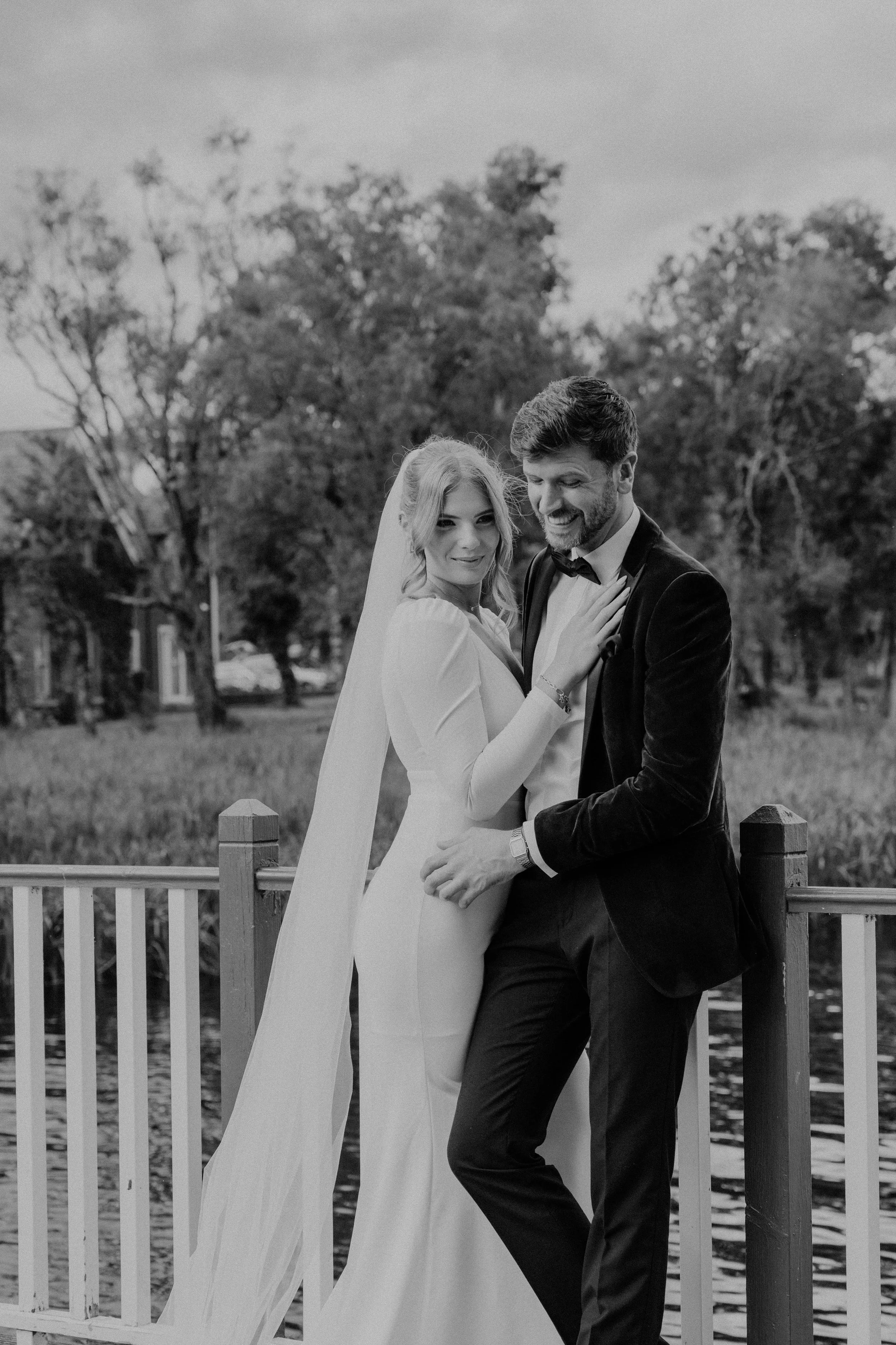 Black and white photo of a bride and groom standing on a bridge by a river, embracing and smiling, with trees in the background.