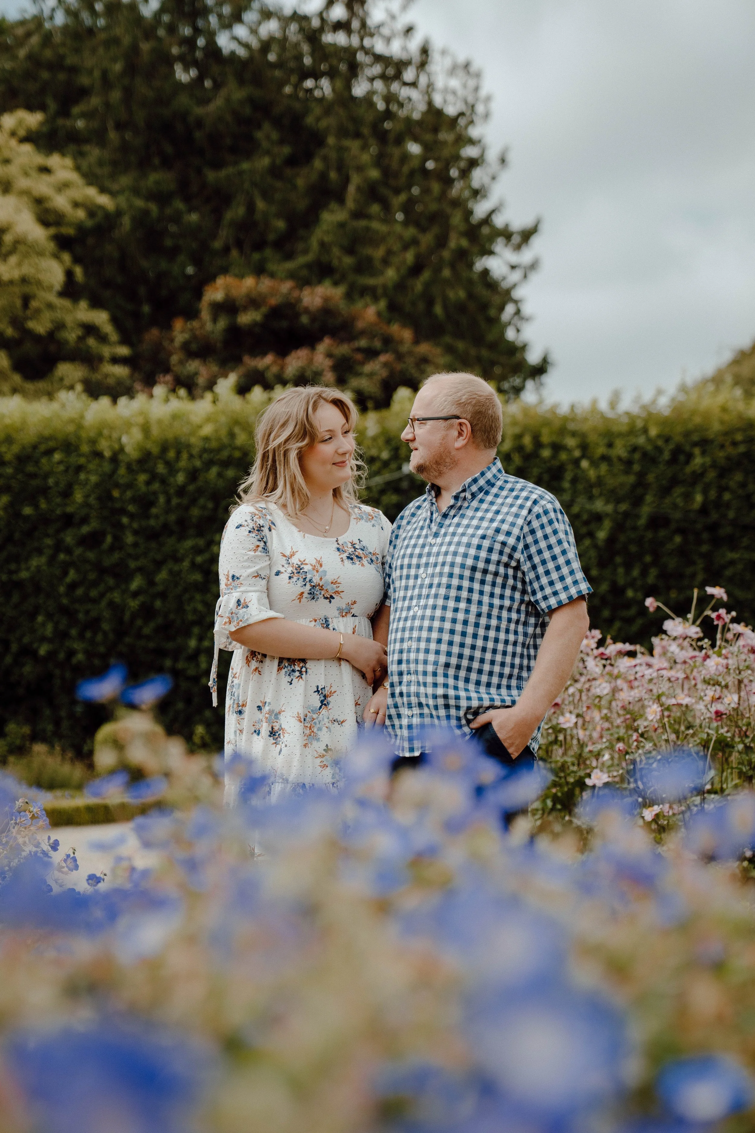 A couple standing close together in a garden with bushes and flowers, looking at each other.