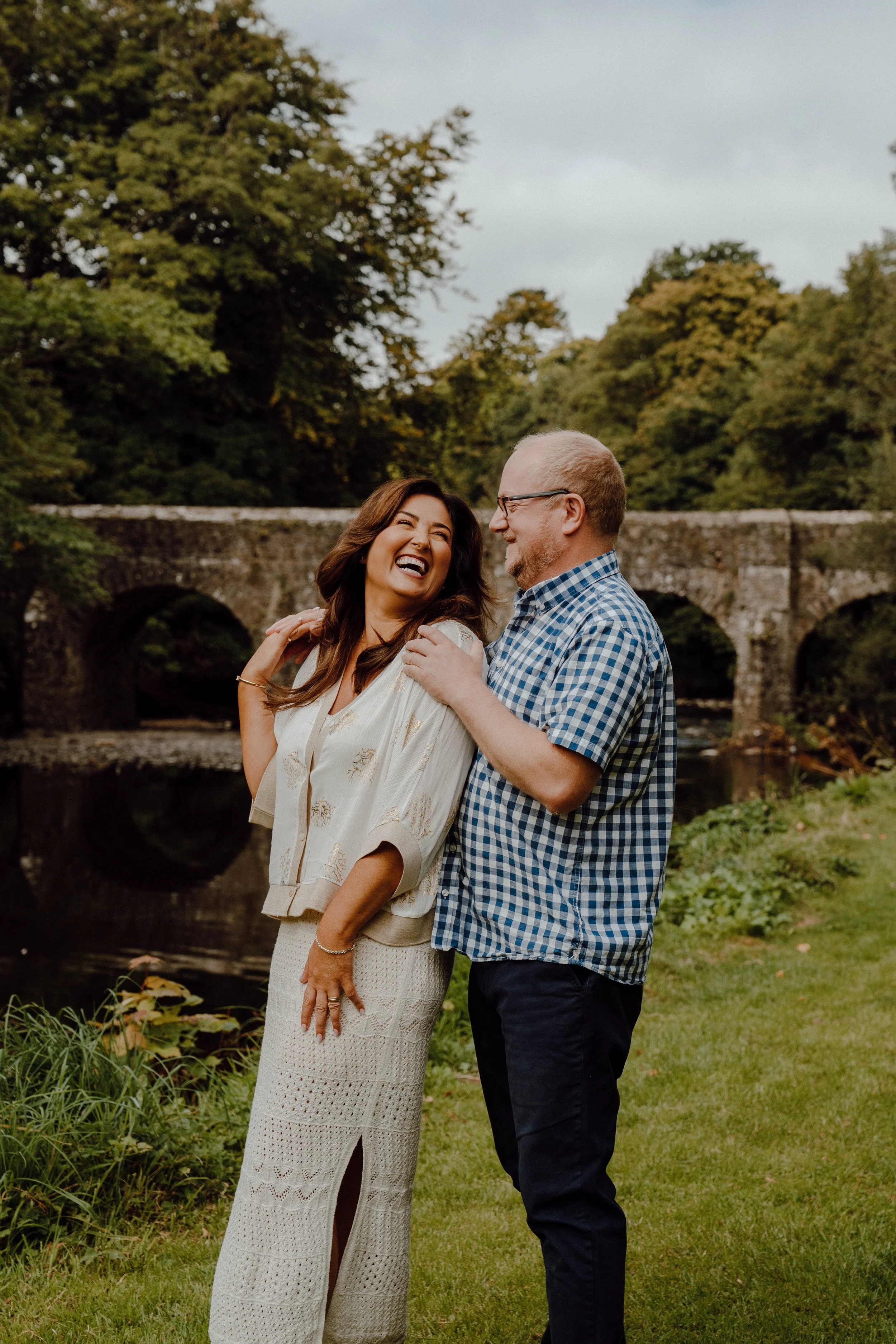 A joyful couple standing outdoors near a river with a stone bridge in the background. They are laughing and embracing each other, surrounded by green trees and grass.